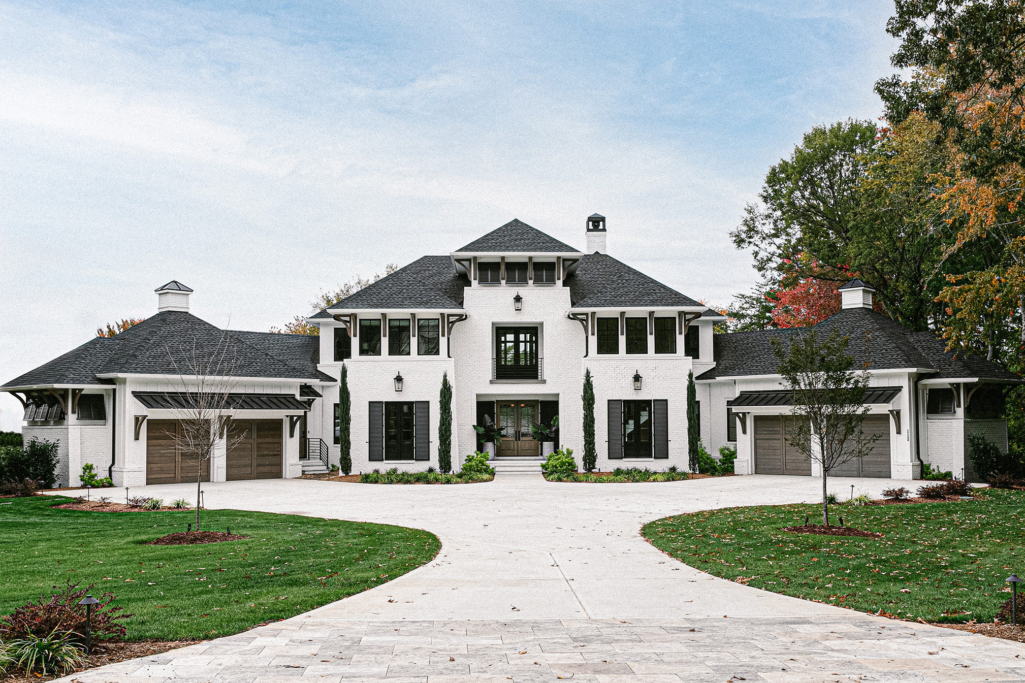 Front exterior of a custom-built white painted brick estate by PJD Homes in Lake Norman featuring a symmetrical façade with dark shutters, stained wood garage doors, Italian cypress landscaping, and a circular paver driveway.