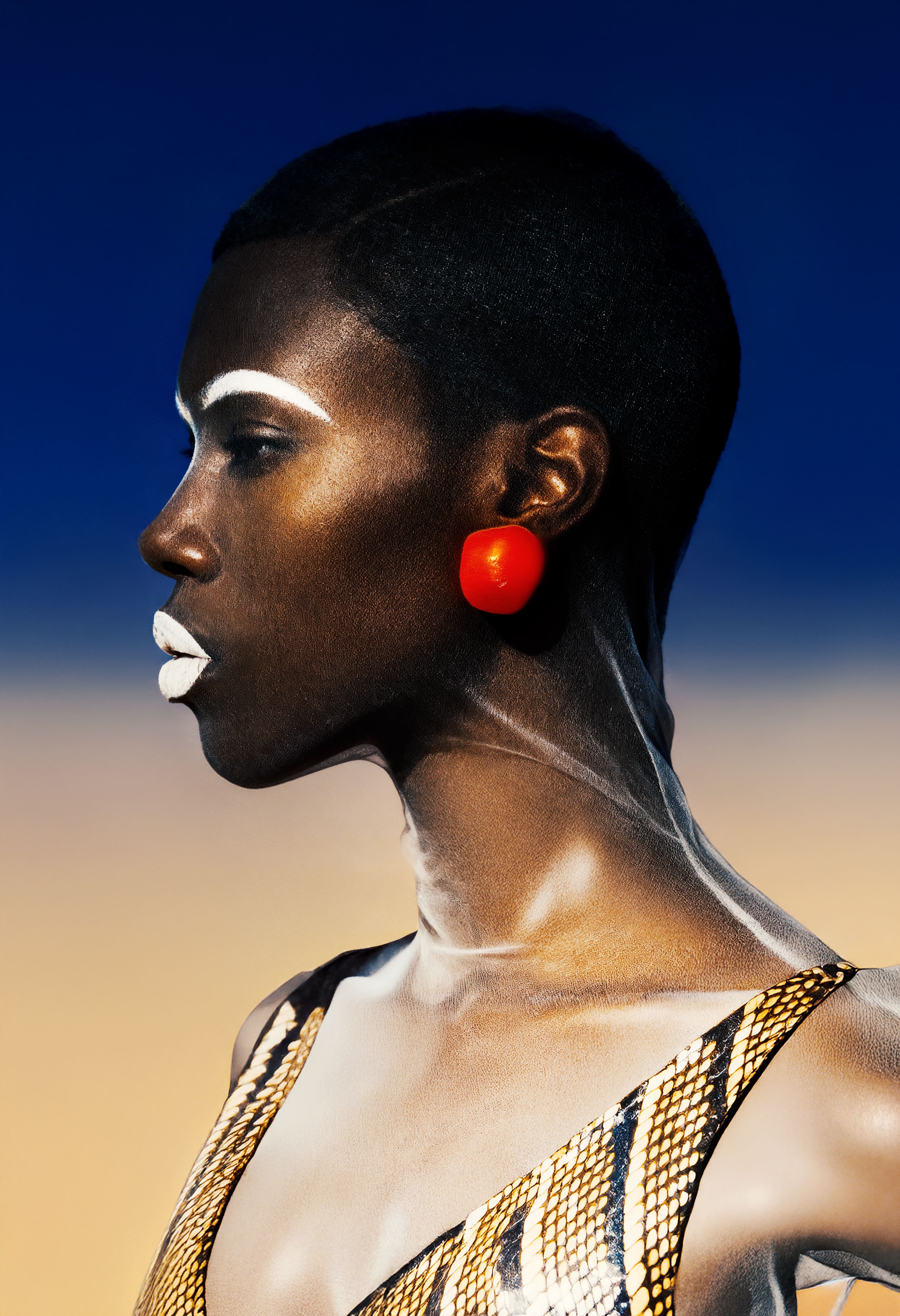 Profile of a woman with short hair, white face paint on eyebrows and lips, red round earrings, and a patterned top against a gradient blue to beige background.