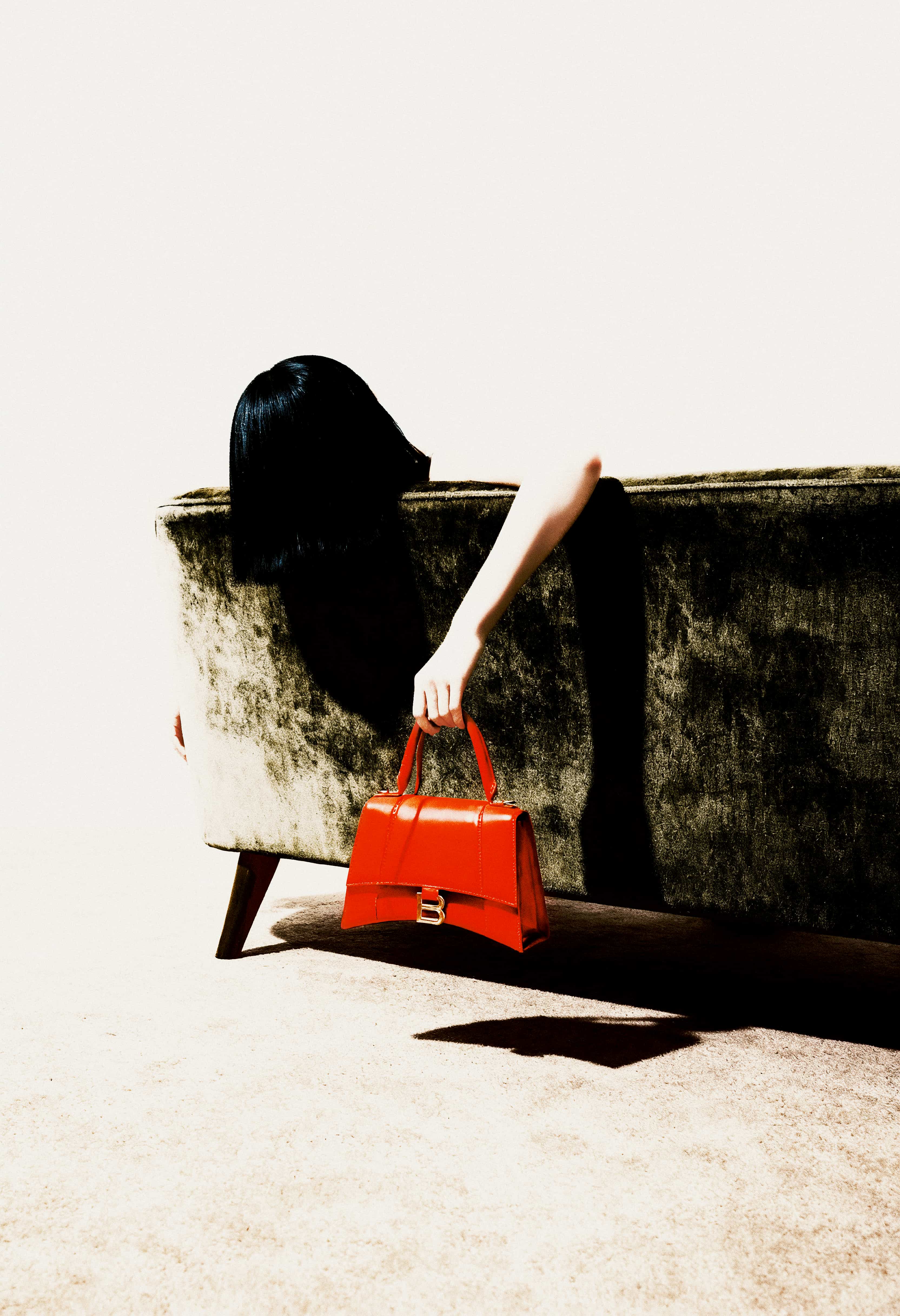 Person with black hair leaning over the back of a dark green velvet sofa, holding a bright red handbag with a gold 'B' buckle.