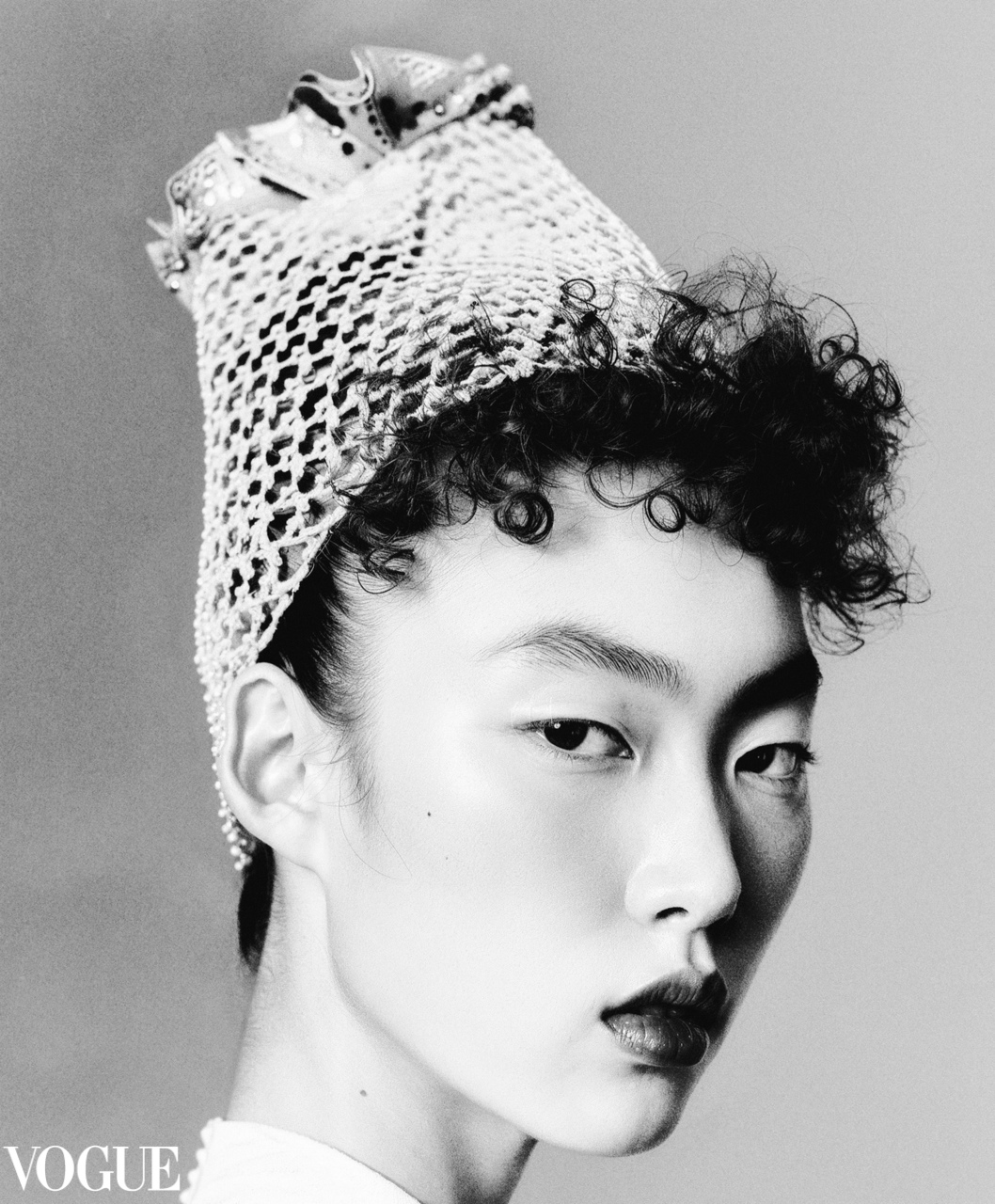 Black and white portrait of a person with short curly hair wearing a textured headpiece, gazing sideways with a serious expression.