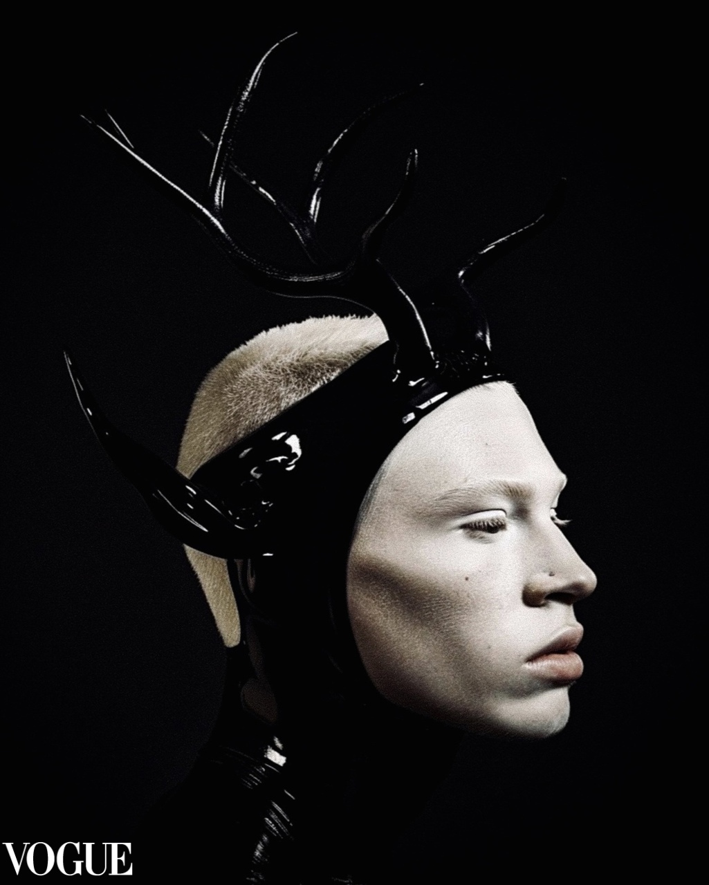Profile of a person with pale skin and short blonde hair wearing a black latex headpiece with glossy black antlers on a dark background.
