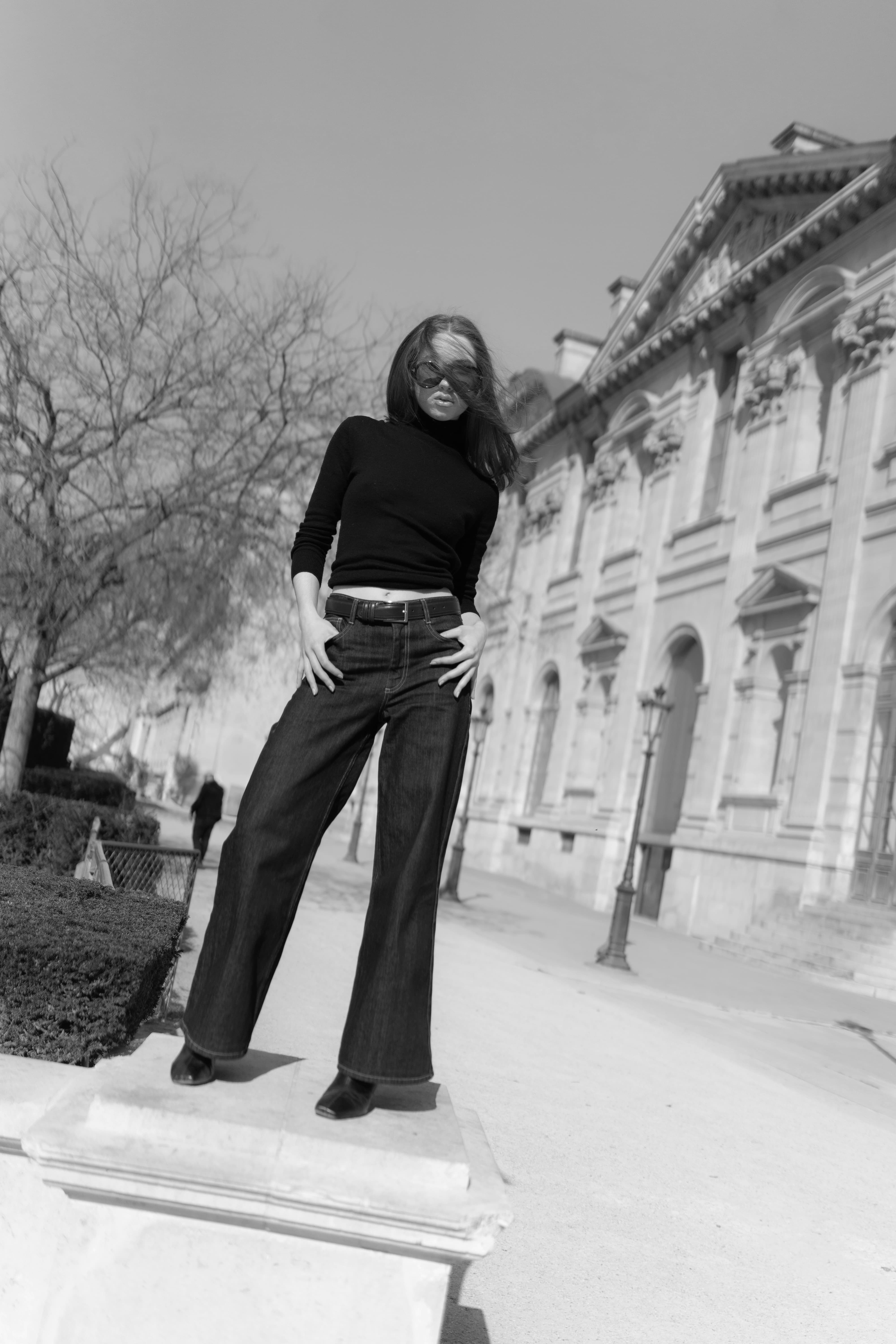 Anna Voytsekhovska in black turtleneck and wide-leg jeans standing on a stone pedestal outdoors near a classical building in Paris.