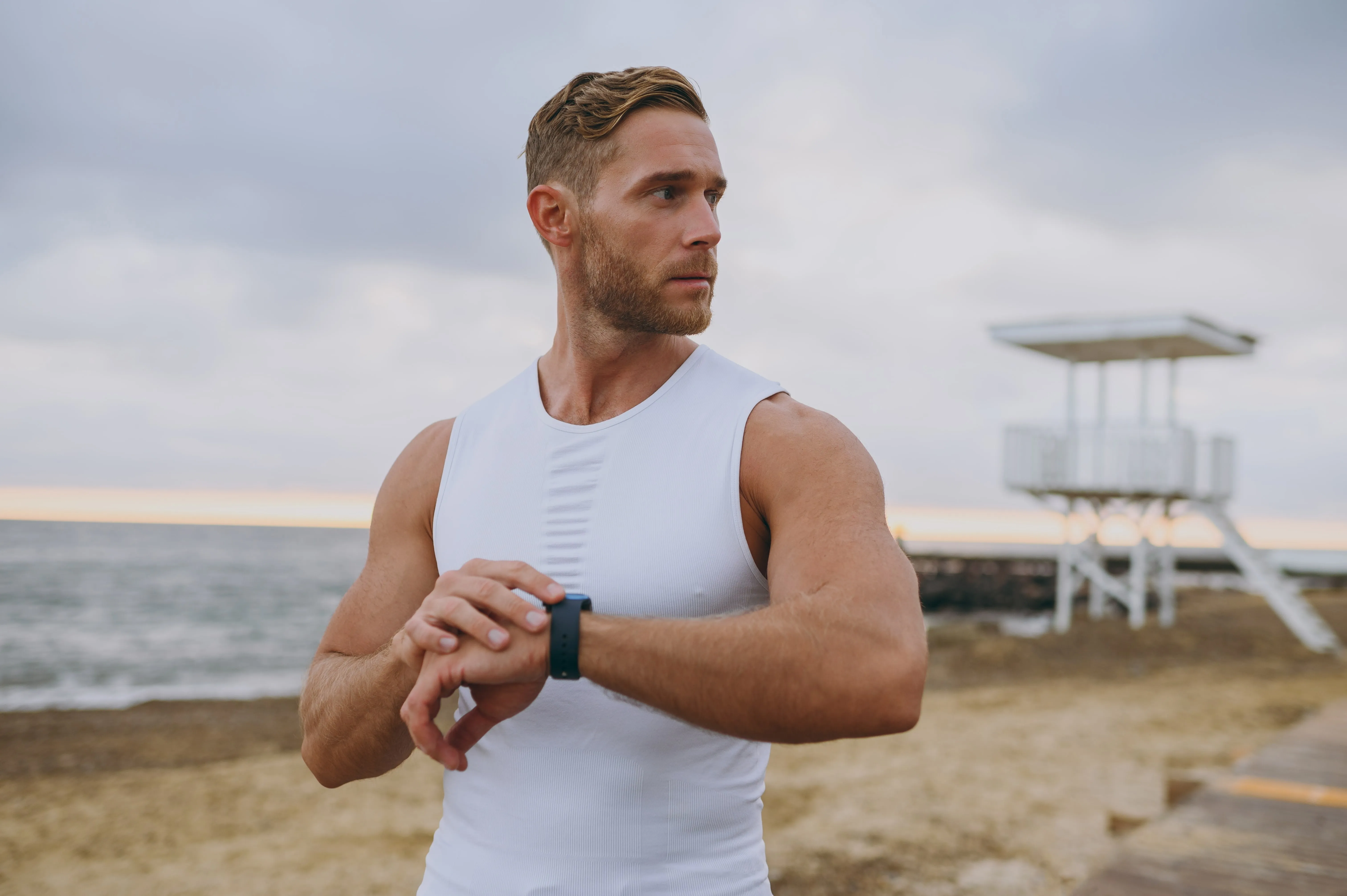 Muscular man in white sleeveless shirt checking a fitness watch on a beach with a lifeguard tower in the background.