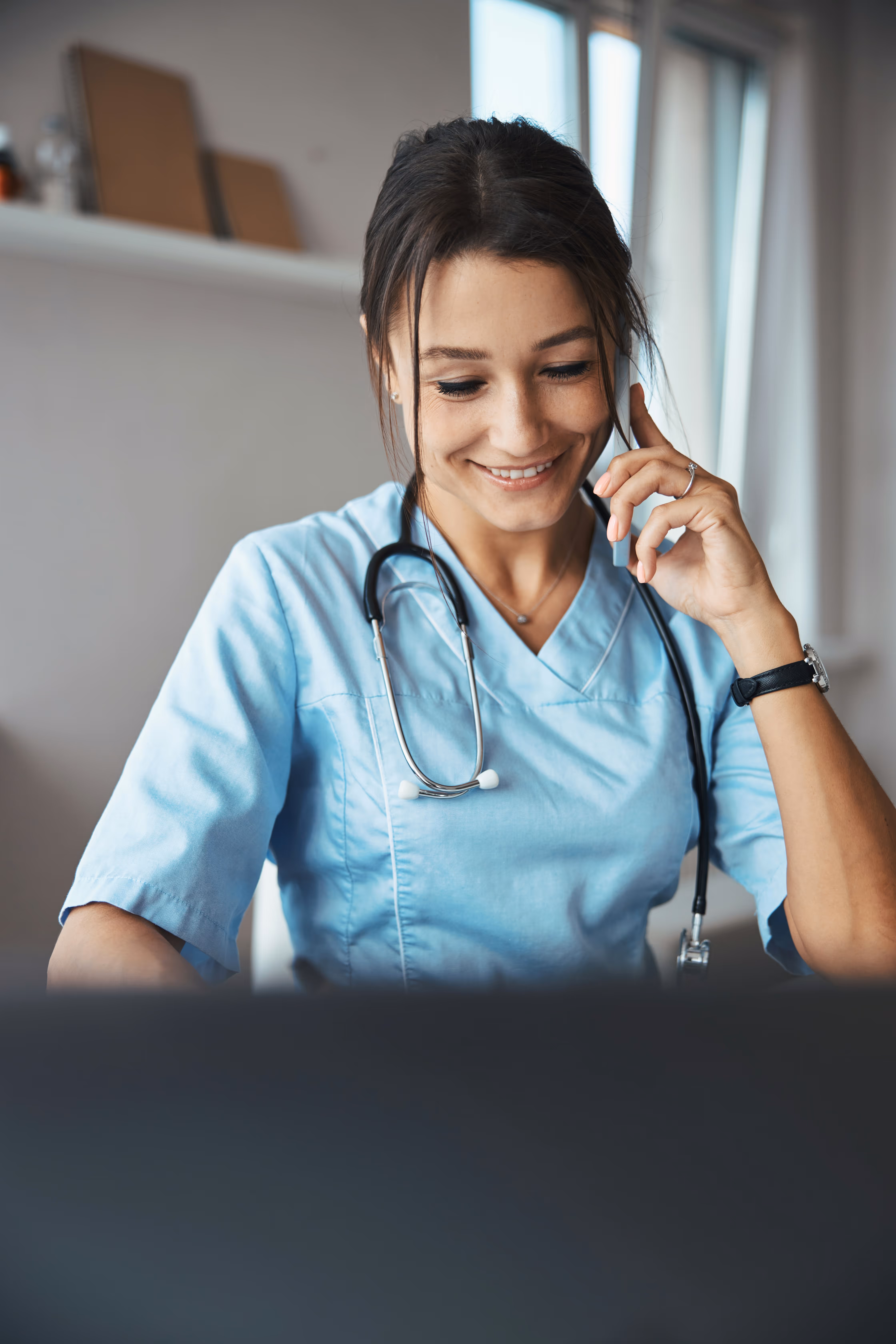 Smiling female doctor in blue scrubs with a stethoscope around her neck, talking on a phone.