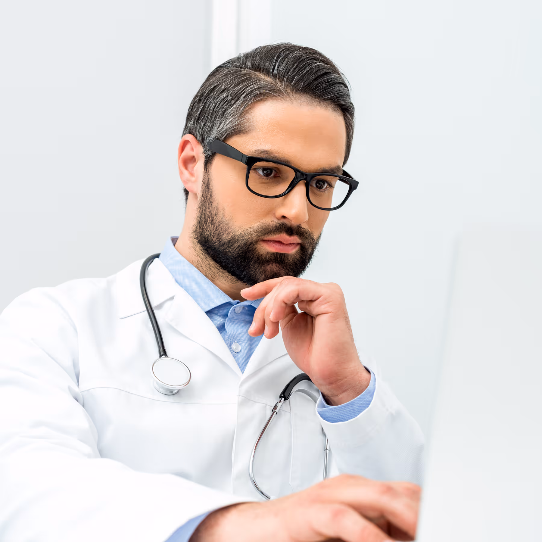 Male doctor wearing white coat, focused on a laptop with hand near his chin.