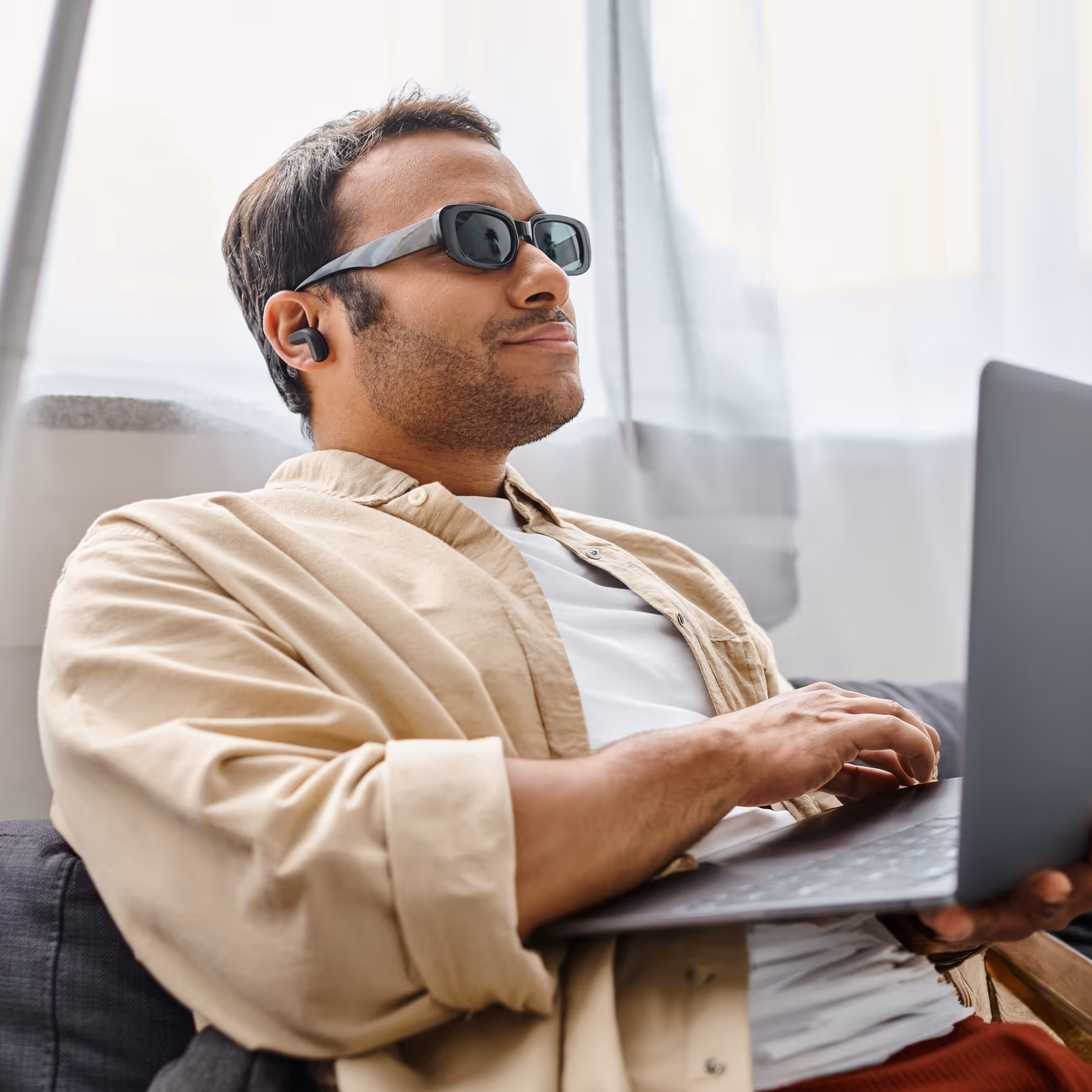 Man wearing sunglasses and earbuds, smiling while using a laptop indoors.