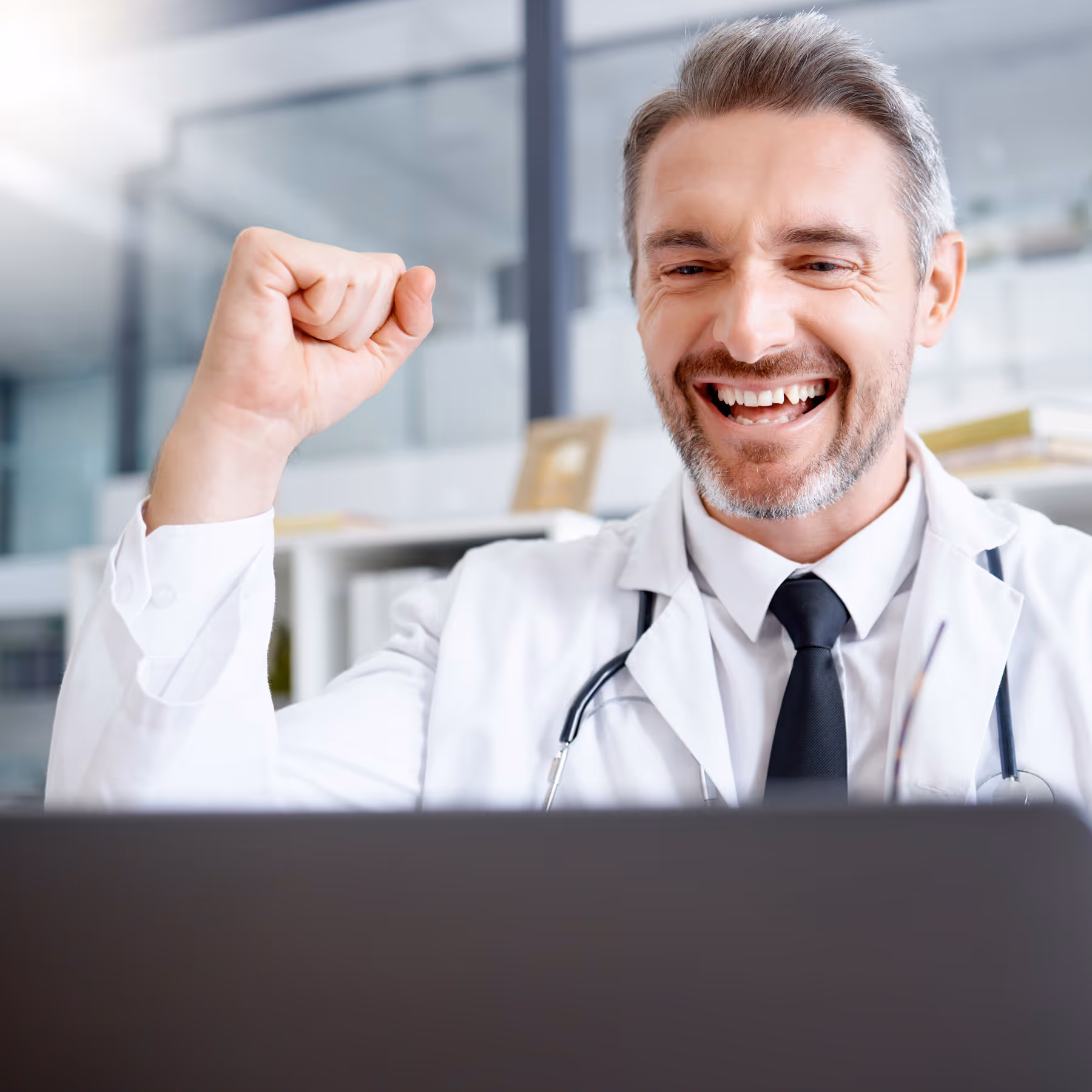 Smiling male doctor in white coat pumping fist with excitement looking at laptop screen.