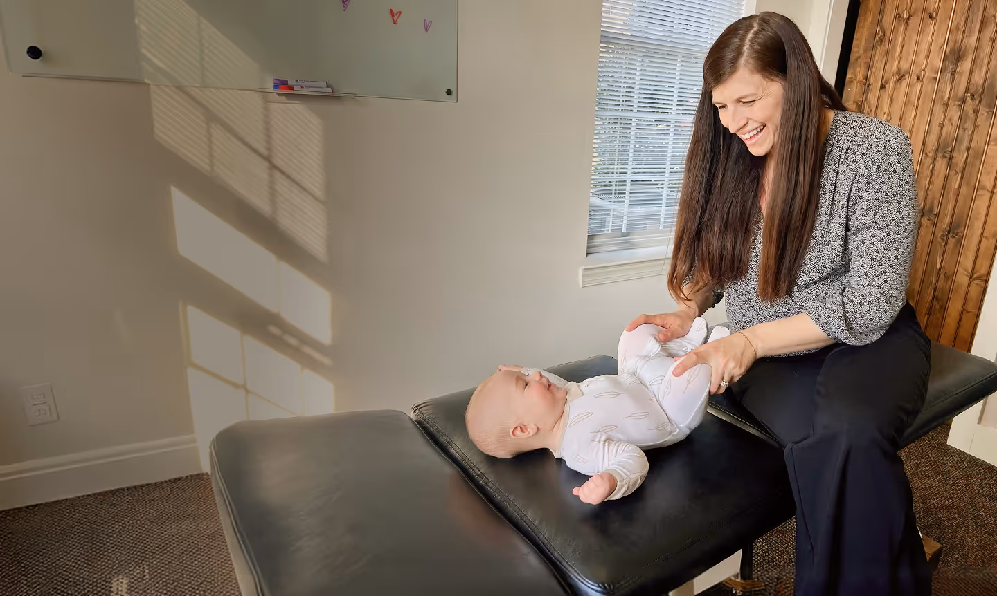 Woman smiling and holding baby's legs while the infant lies on a black padded table.