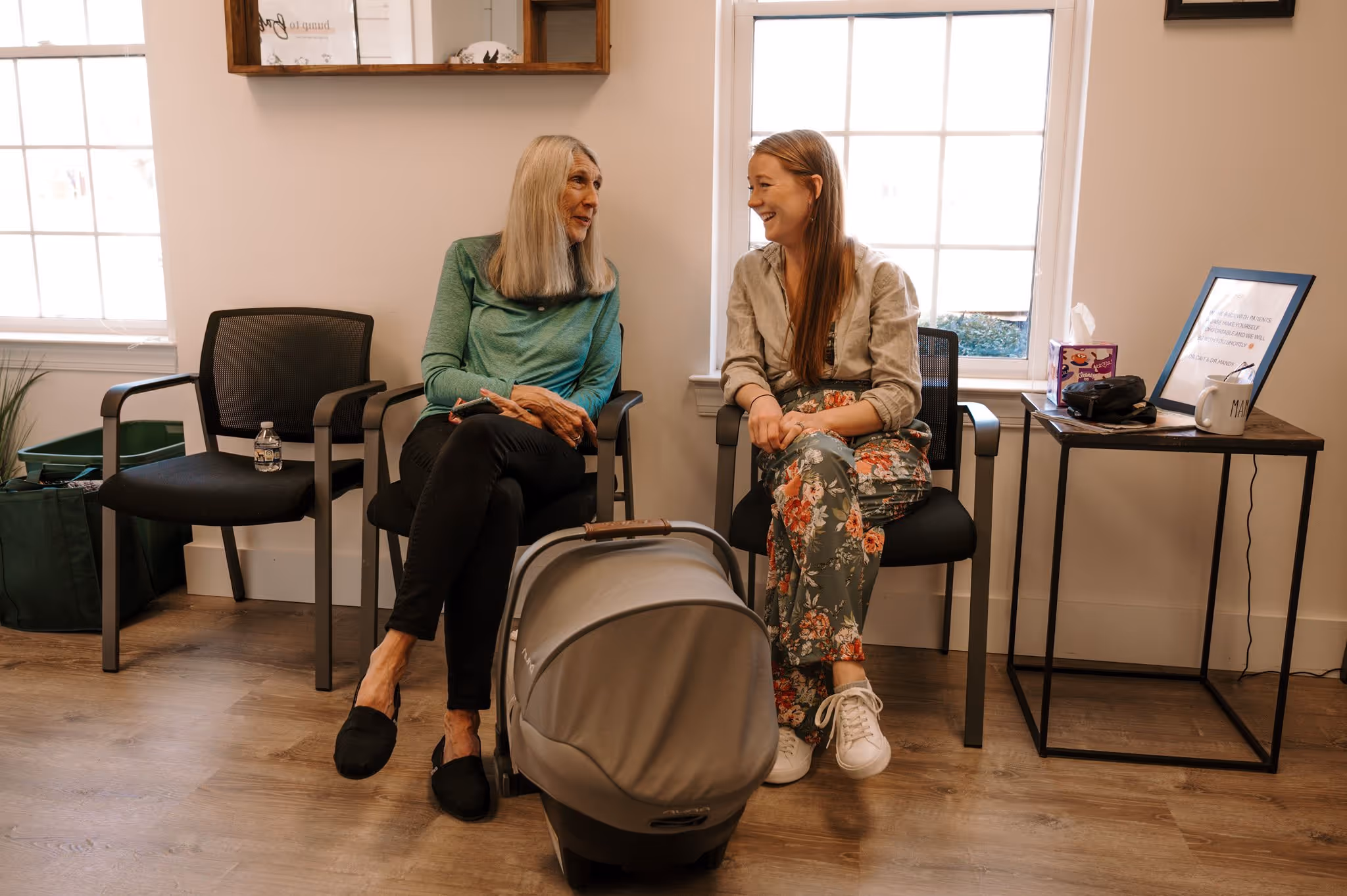 Two women sitting and smiling indoors with a baby car seat in front of them.