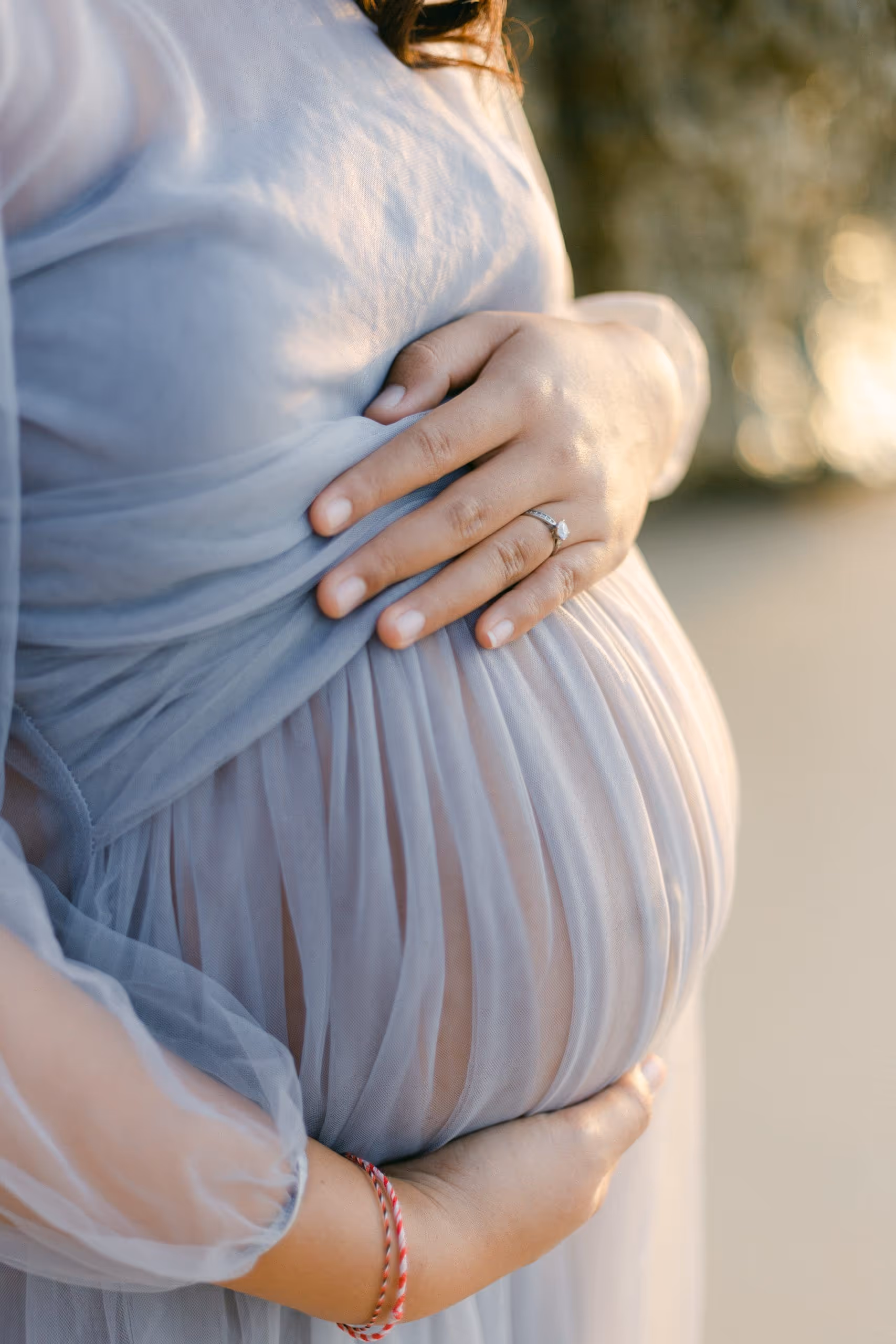 Close-up of pregnant woman in light blue dress holding her belly with both hands outdoors.