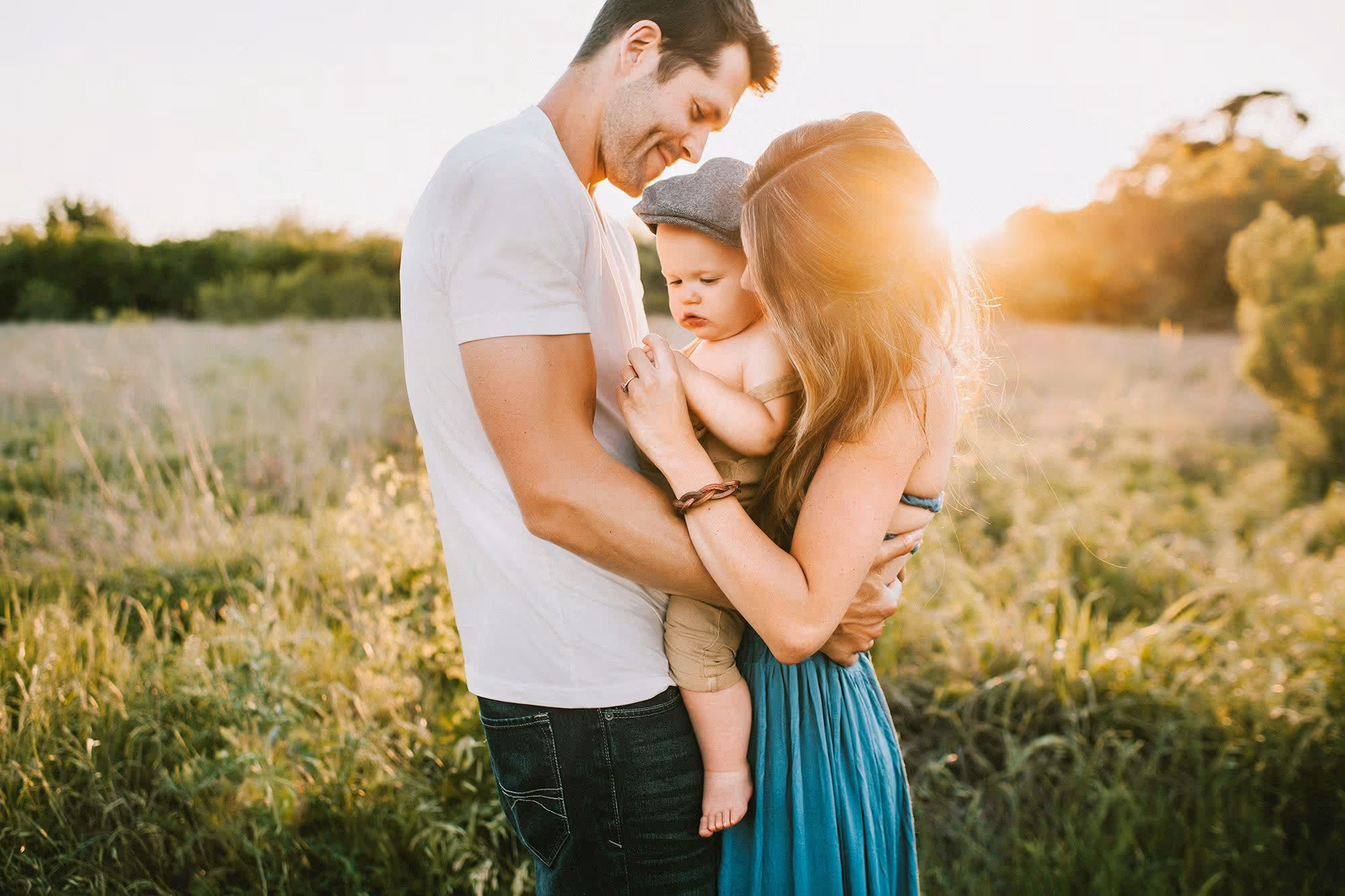 Parents holding and looking at their baby in a sunlit field during golden hour.