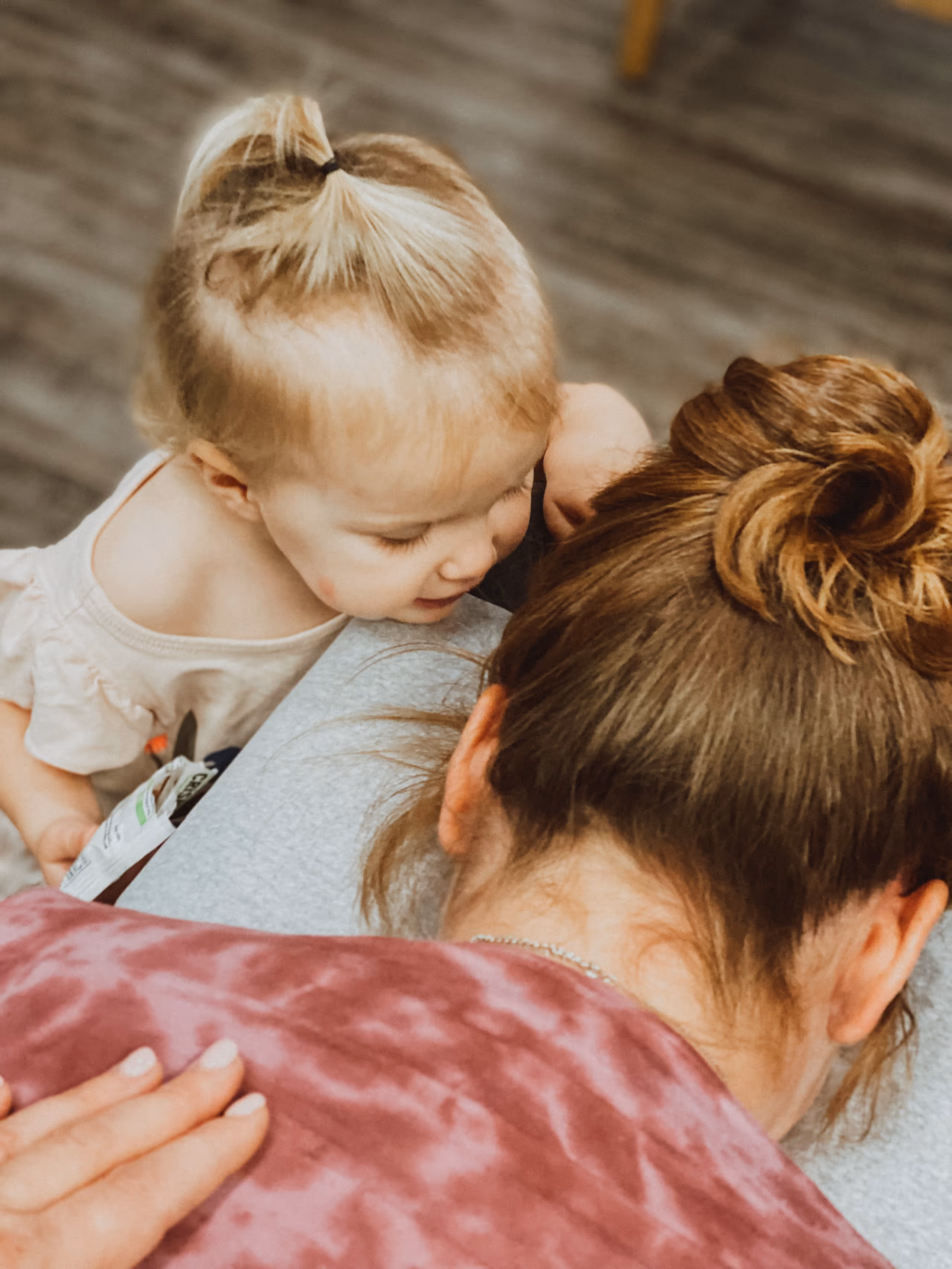 Young child leaning on and embracing adult with hair in a bun, indoors on wooden floor.