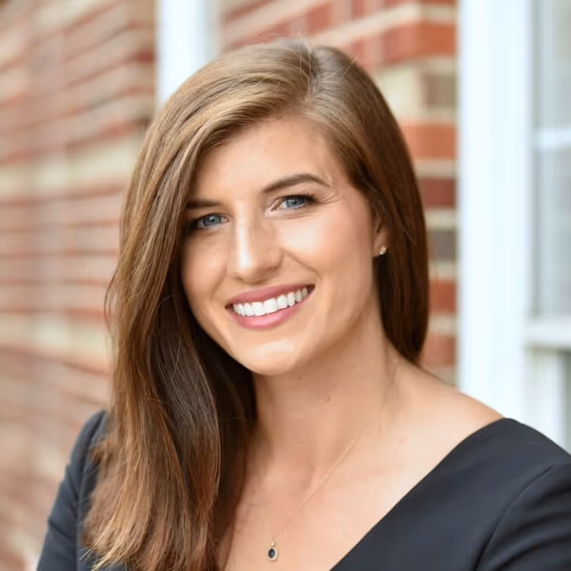 Smiling woman with long brown hair and blue eyes, wearing a black top and necklace, standing outdoors.