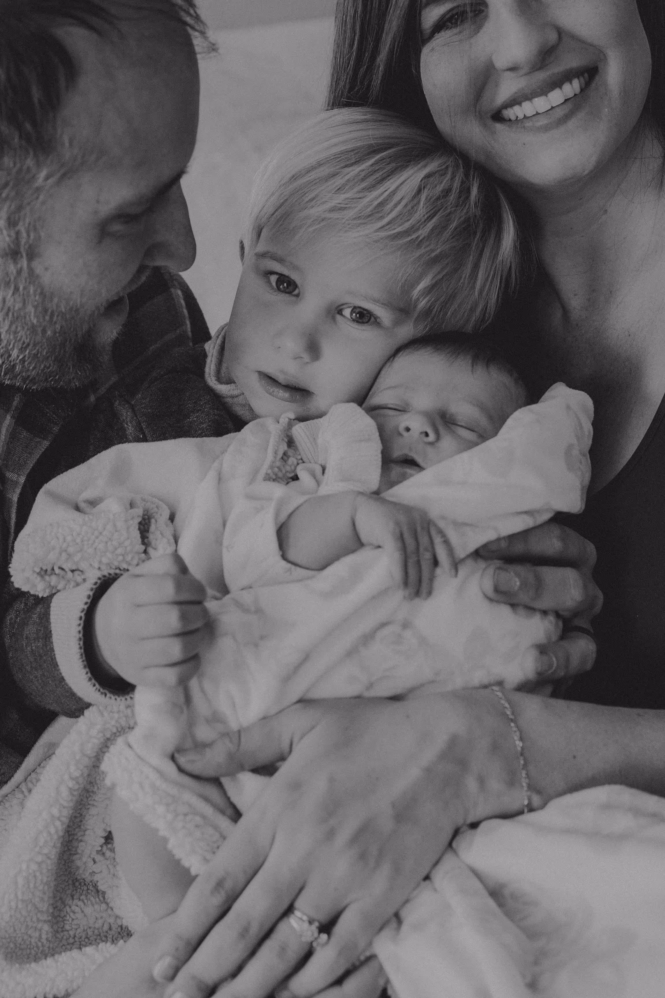 Black and white photo of a smiling family with a toddler holding a sleeping newborn wrapped in a blanket.