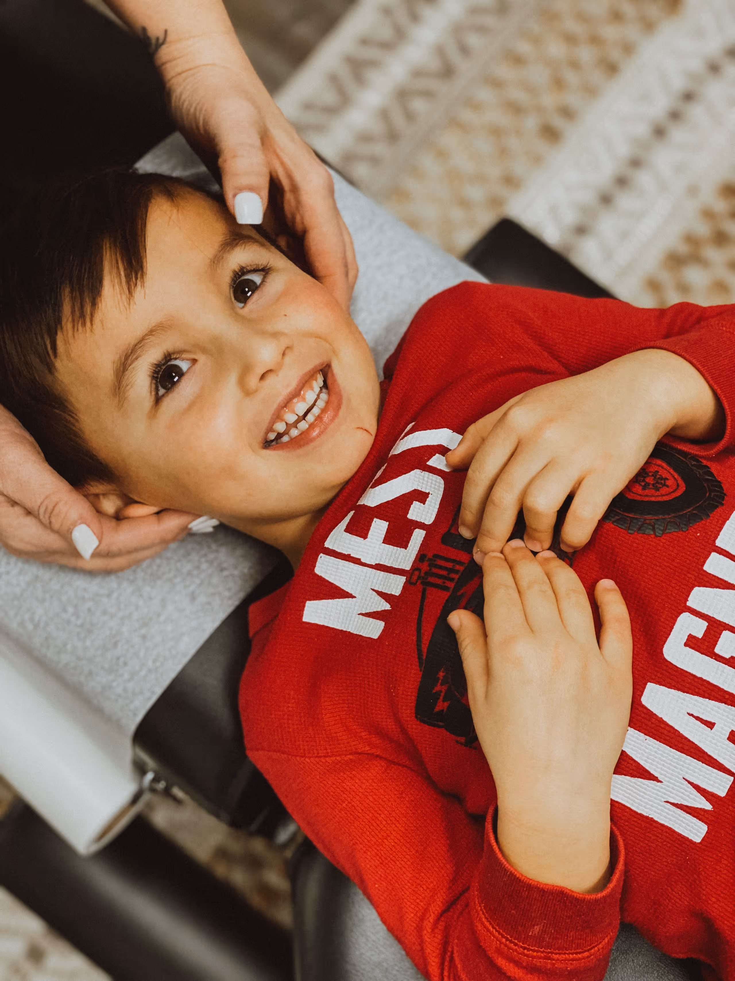 Smiling boy in red shirt lying down while an adult gently holds his head with painted nails.