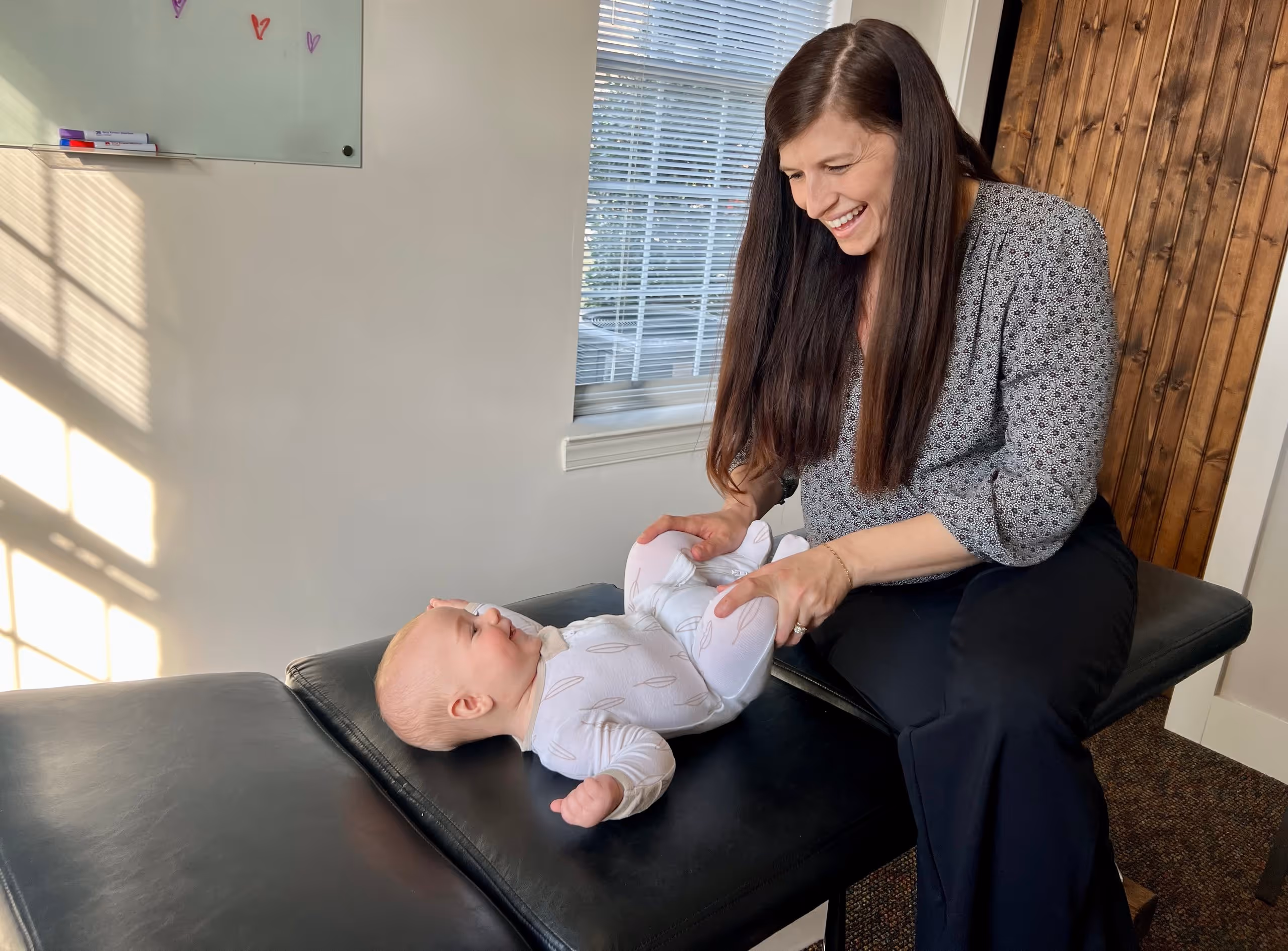 Woman smiling and holding legs of a baby lying on a black chiropractor's table.