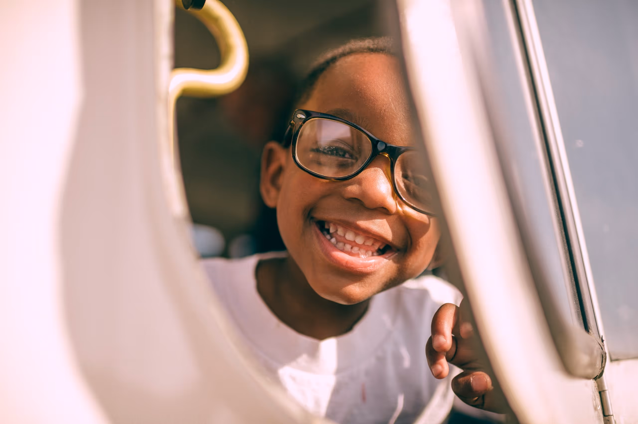 Smiling child wearing glasses peeks through a window, holding onto the edge with one hand.