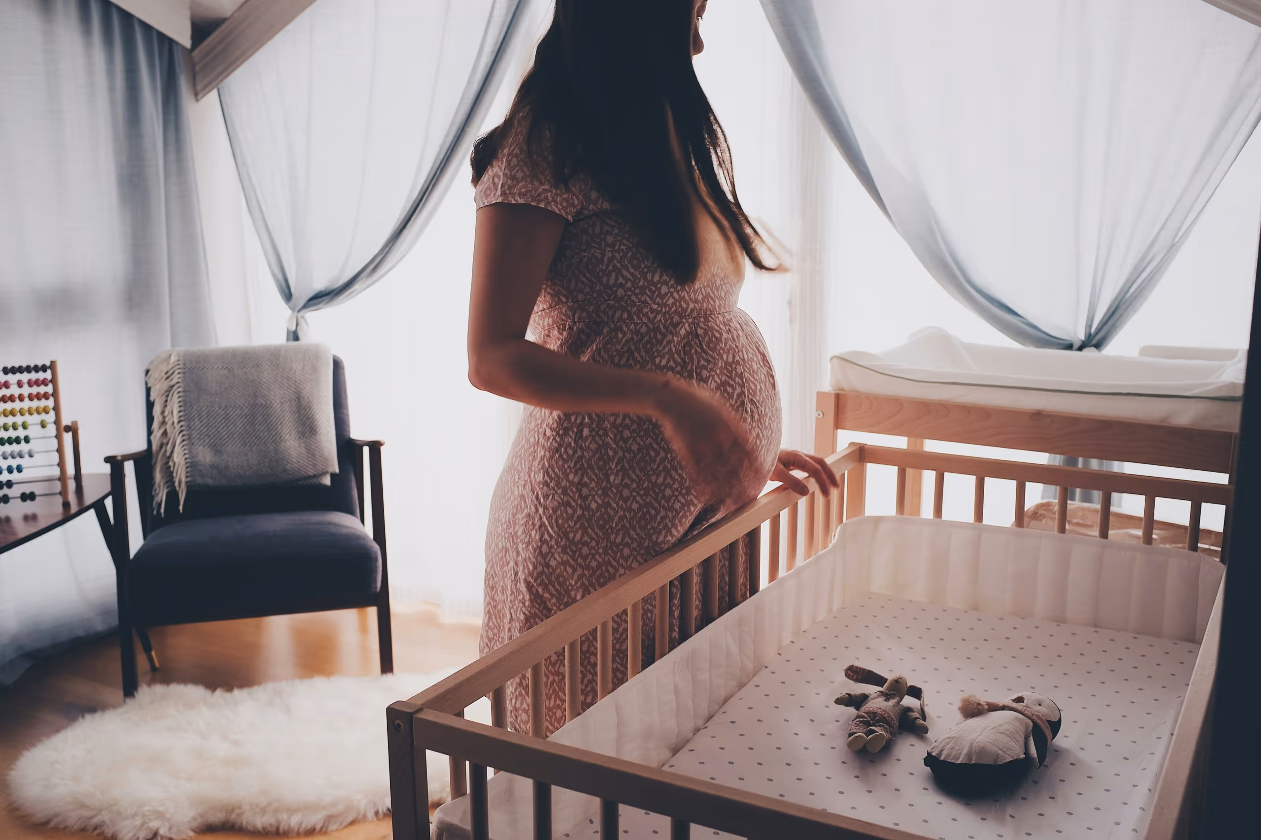 Pregnant woman standing by a wooden baby crib with two stuffed toys in softly lit nursery.