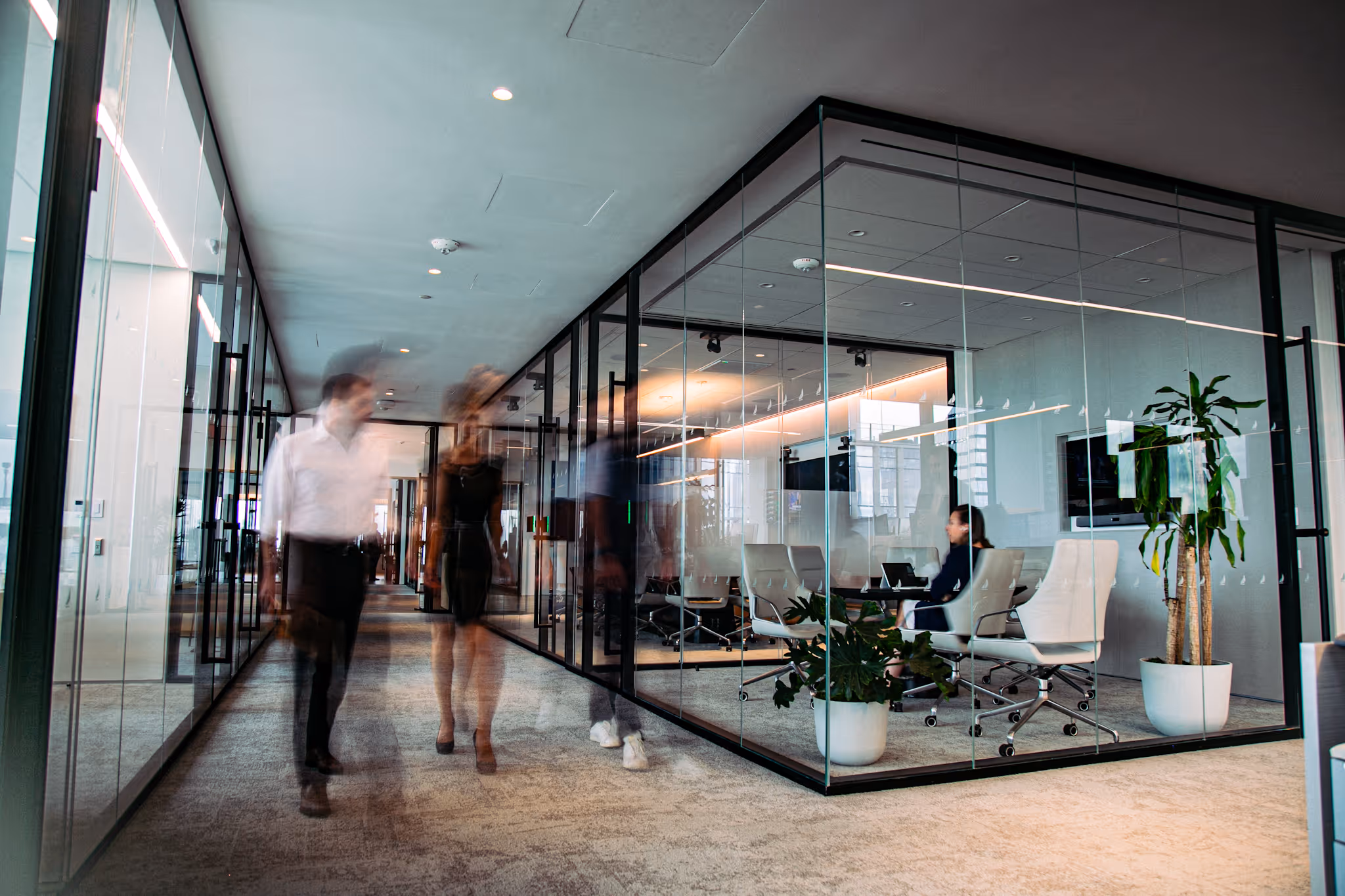 Modern office corridor with glass-walled conference room on the right and blurred figures walking in the hallway.