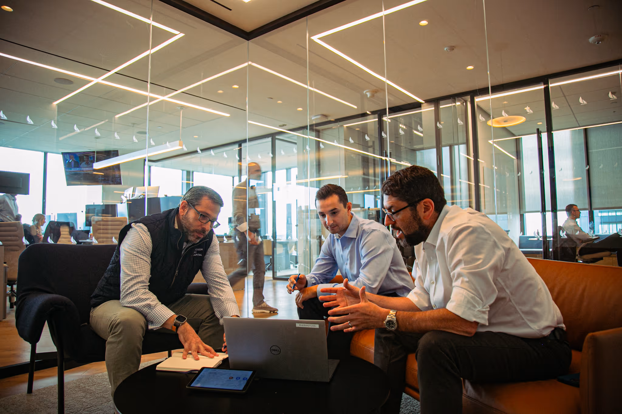 Three men engaged in a discussion around a laptop and tablet in a modern glass-walled office meeting area.