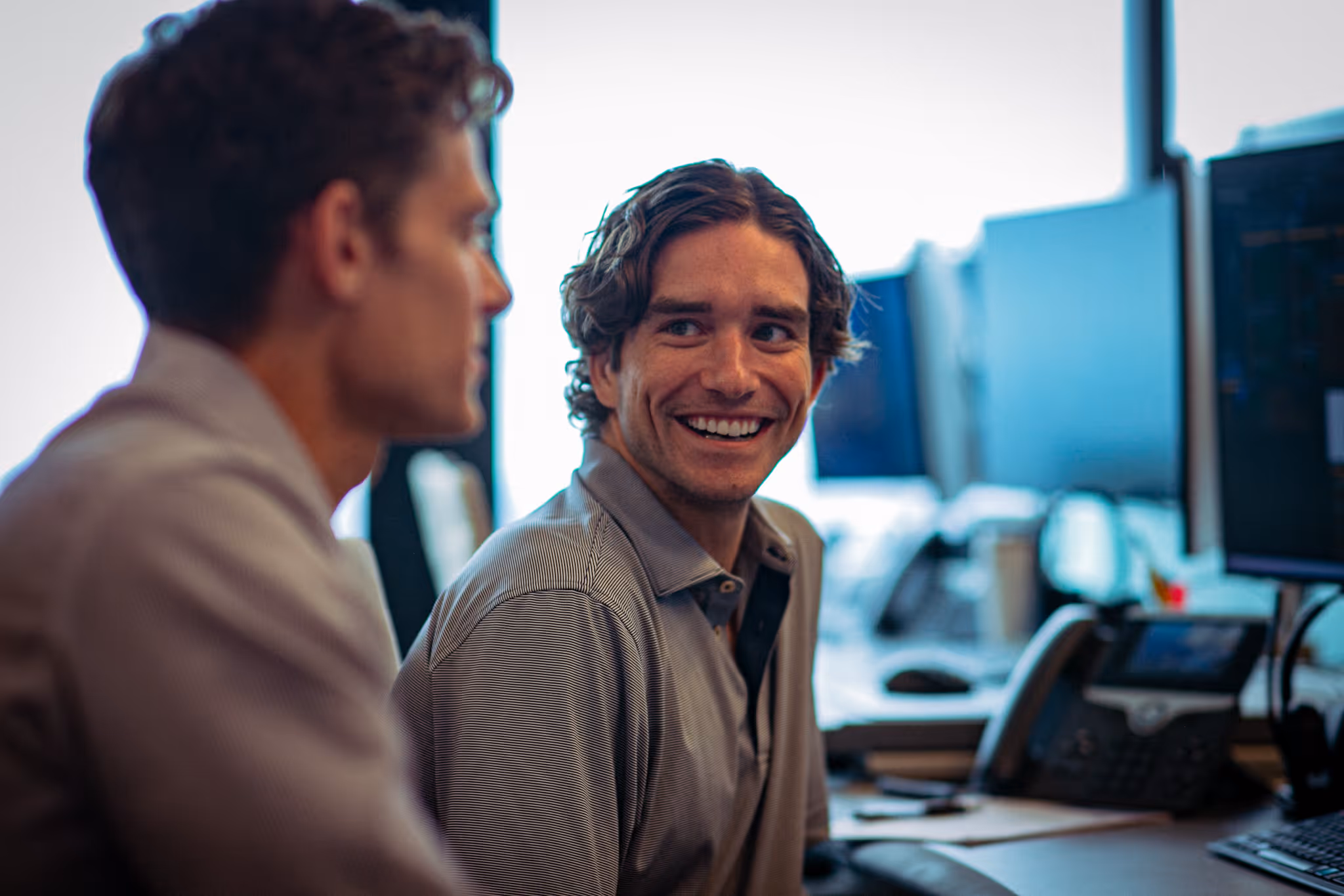 Two young men in an office setting, one smiling at the other while seated near computer monitors and a desk phone.