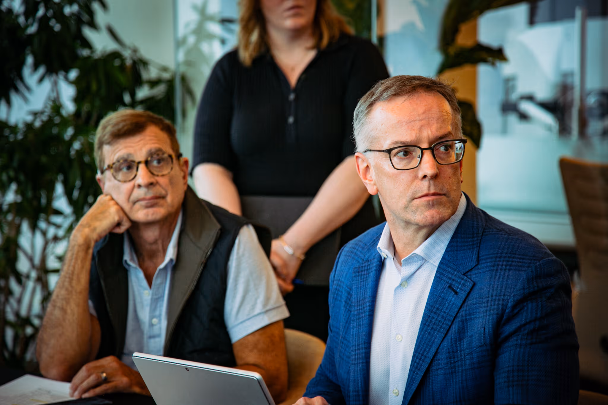 Three men engaged in a discussion around a laptop and tablet in a modern glass-walled office meeting area.