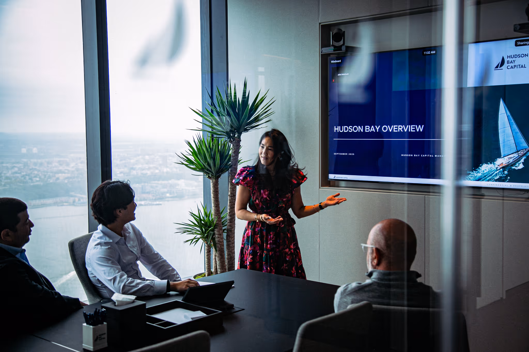 Woman presenting a business overview titled 'Hudson Bay Overview' to three colleagues in a modern conference room with a large window view.