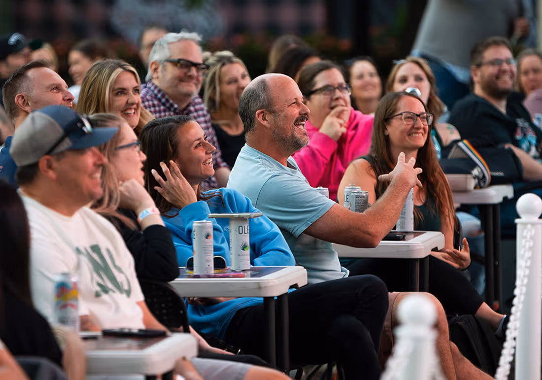 Group of people sitting closely at a comendy festival, smiling and laughing during an outdoor event.