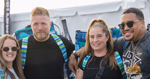 Four smiling adults standing closely outdoors at a comedy festival with backpacks and sunglasses.