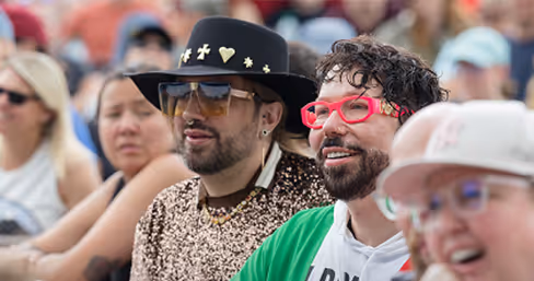 Two men sitting among a comedy crowd, one wearing a black hat and sunglasses, the other red glasses and green jacket.