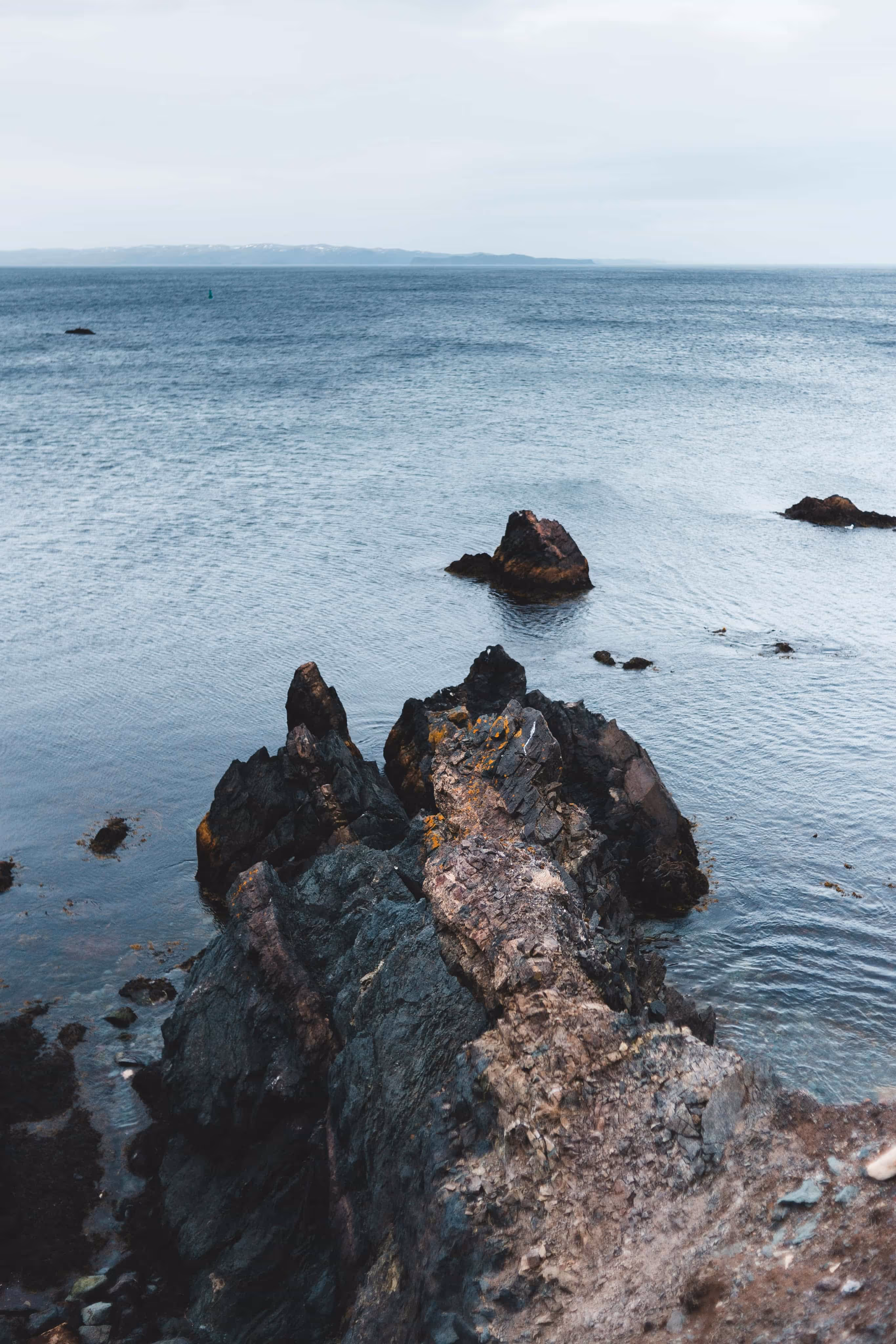 Jagged dark rocky outcrop extending into calm blue ocean under a cloudy sky.