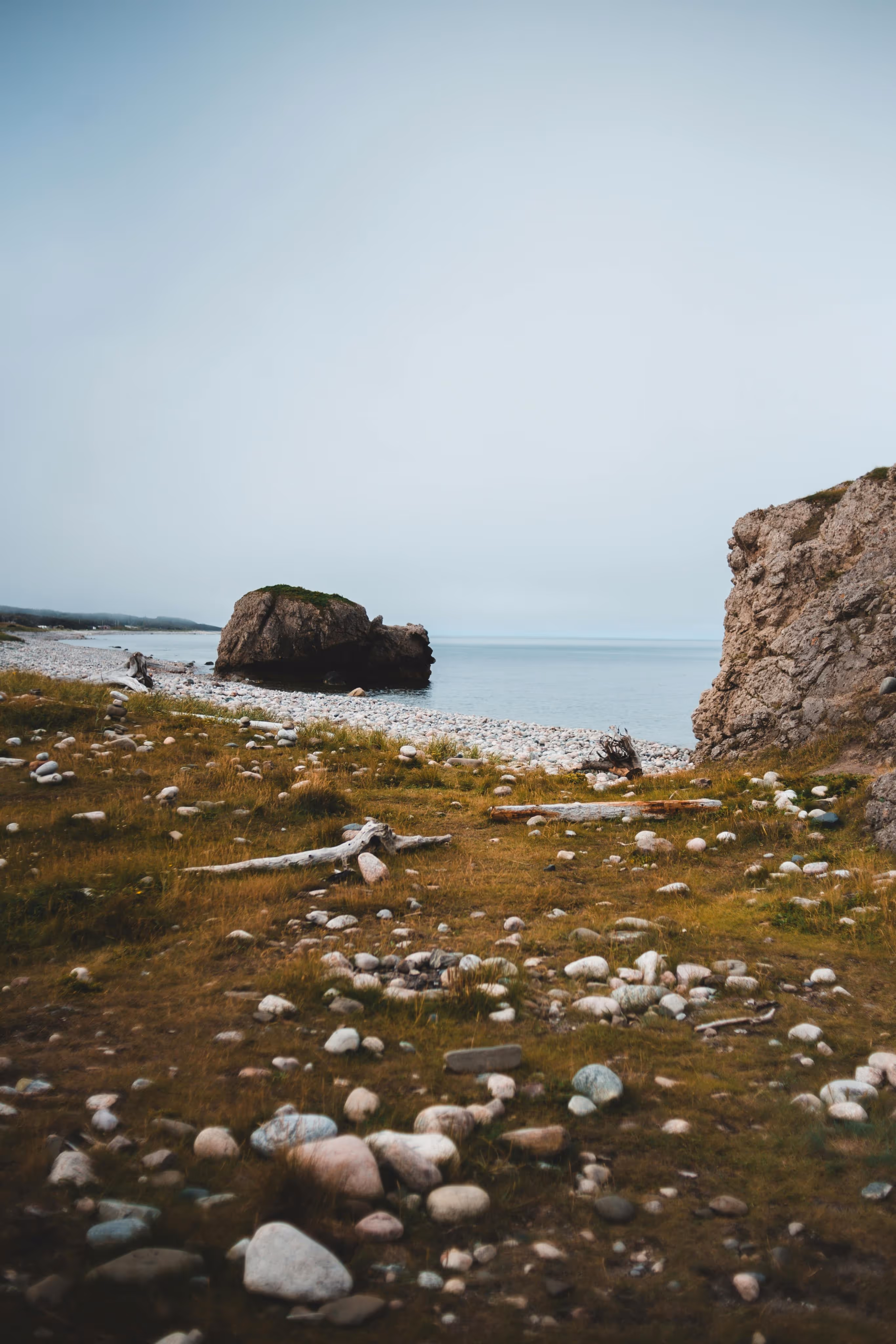 Rocky shoreline with scattered white stones, driftwood, and large rock formations by calm sea under a cloudy sky.