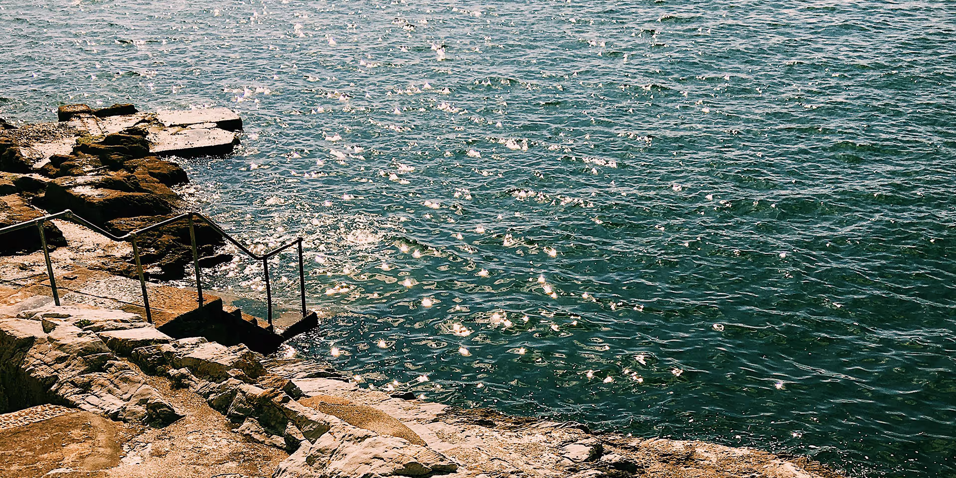 Sunlit rocky shoreline with metal handrails leading down to the water.