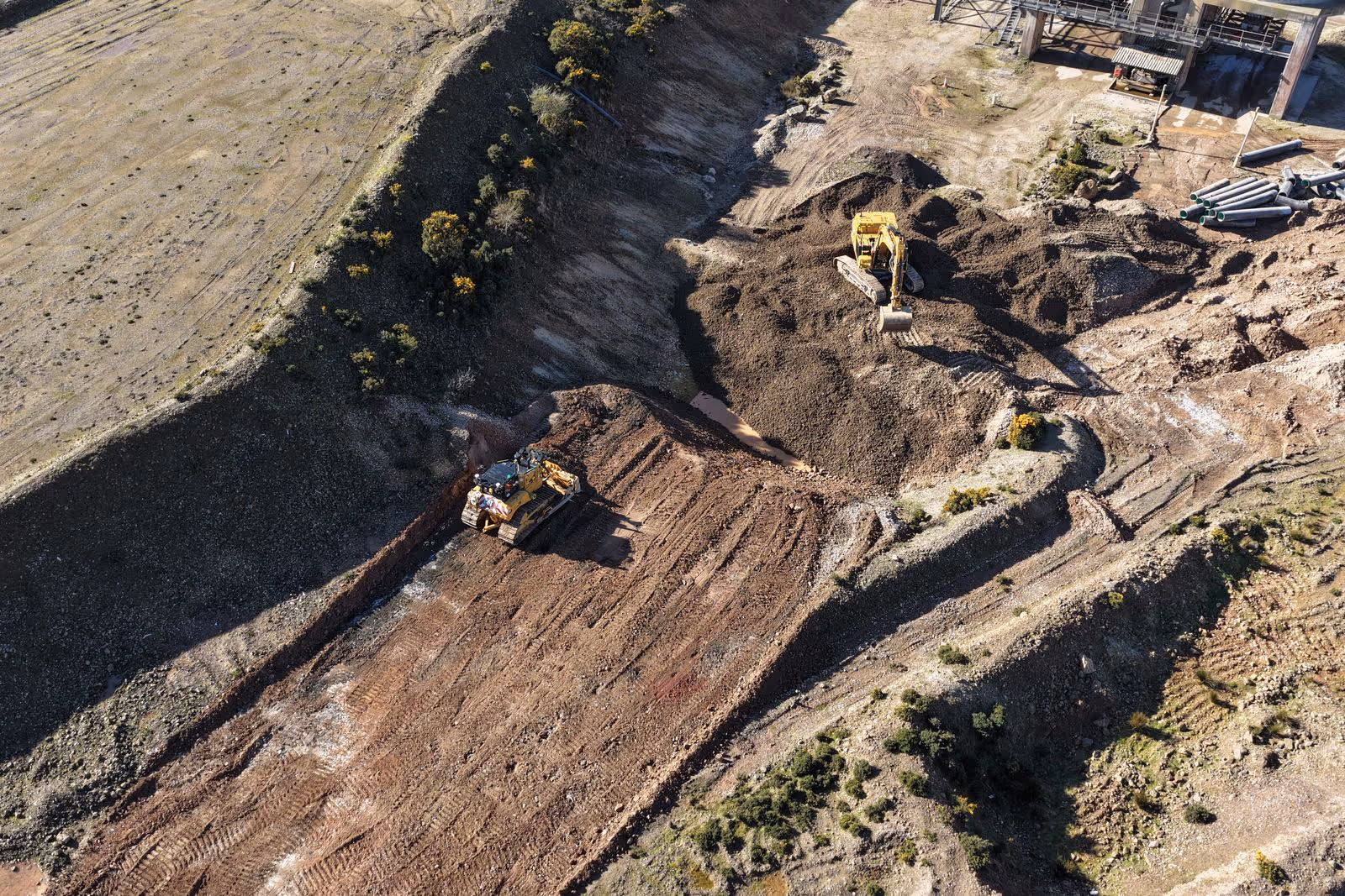 Aerial view of excavators and bulldozer working on a construction site with piles of dirt and rugged terrain.