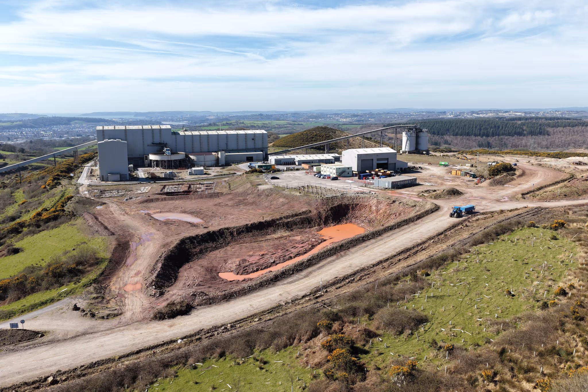 Aerial view of an industrial mining site with large buildings, dirt roads, excavation pits filled with reddish water, and surrounding green landscape.