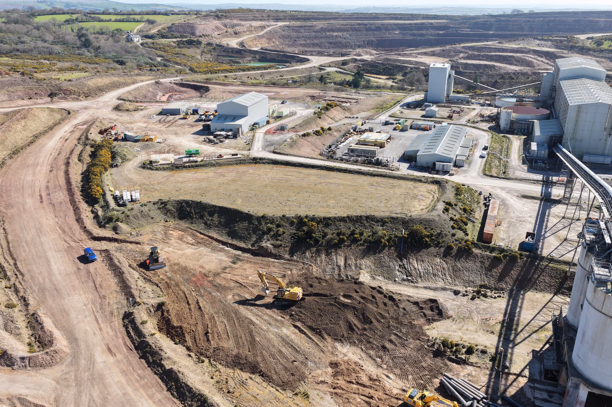 Aerial view of an industrial quarry site with excavators, storage buildings, dirt roads, and large open mining areas.