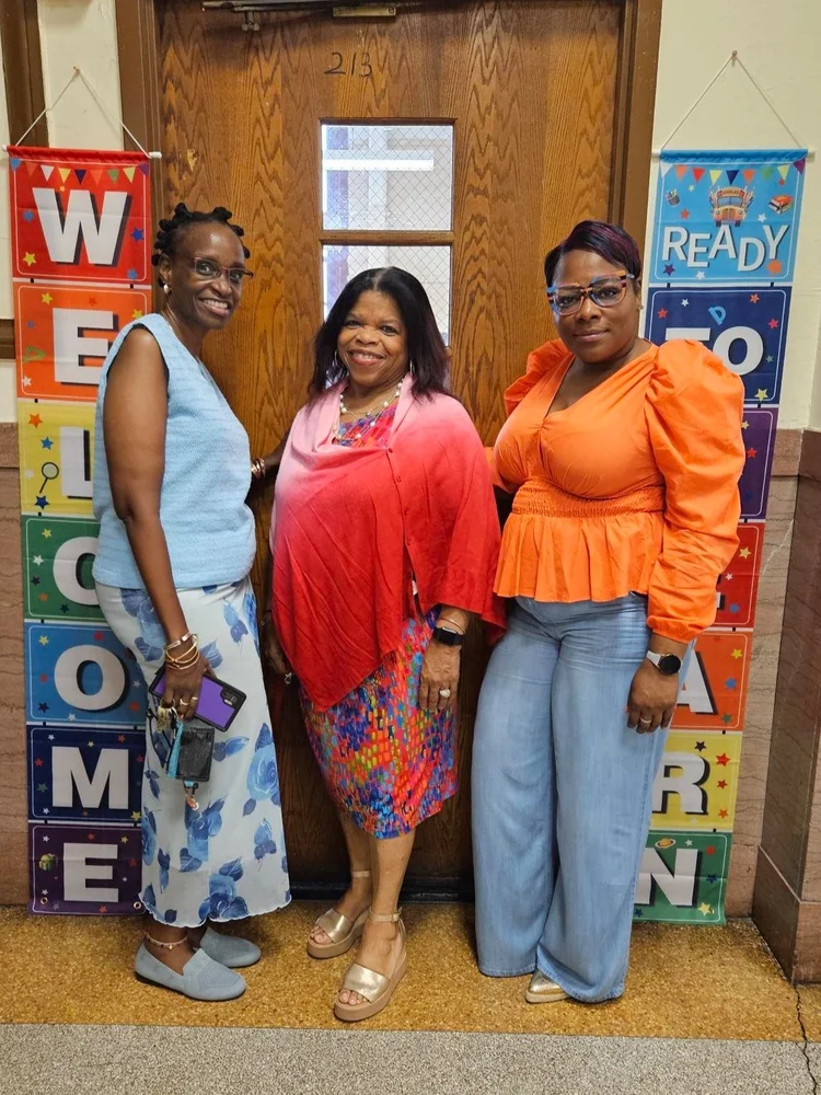 Three staff members smiling outside classroom 213 welcome banners