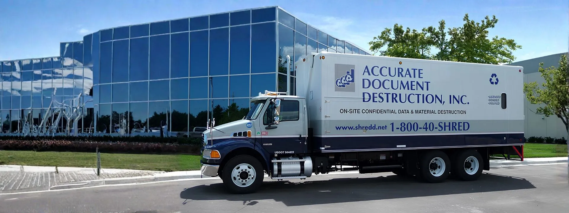 White and blue mobile shredding truck parked in front of a modern glass office building.