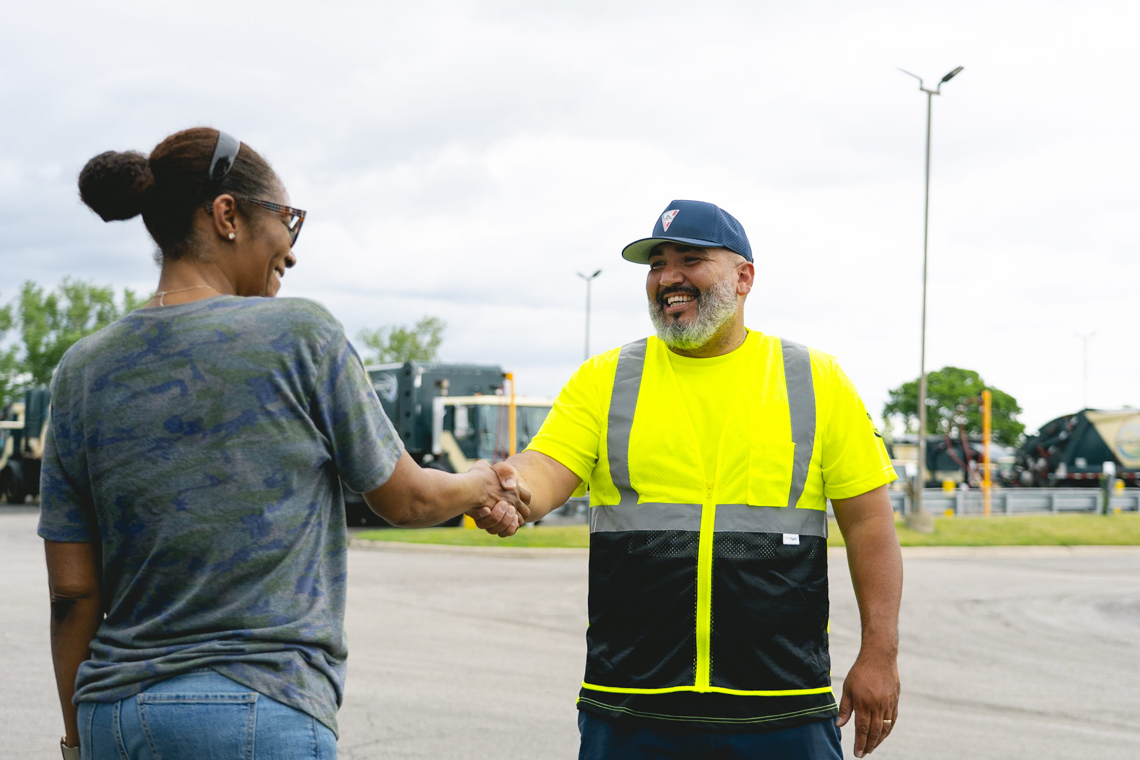 Smiling man in reflective vest shakes hands with woman in casual clothes outdoors near trucks.
