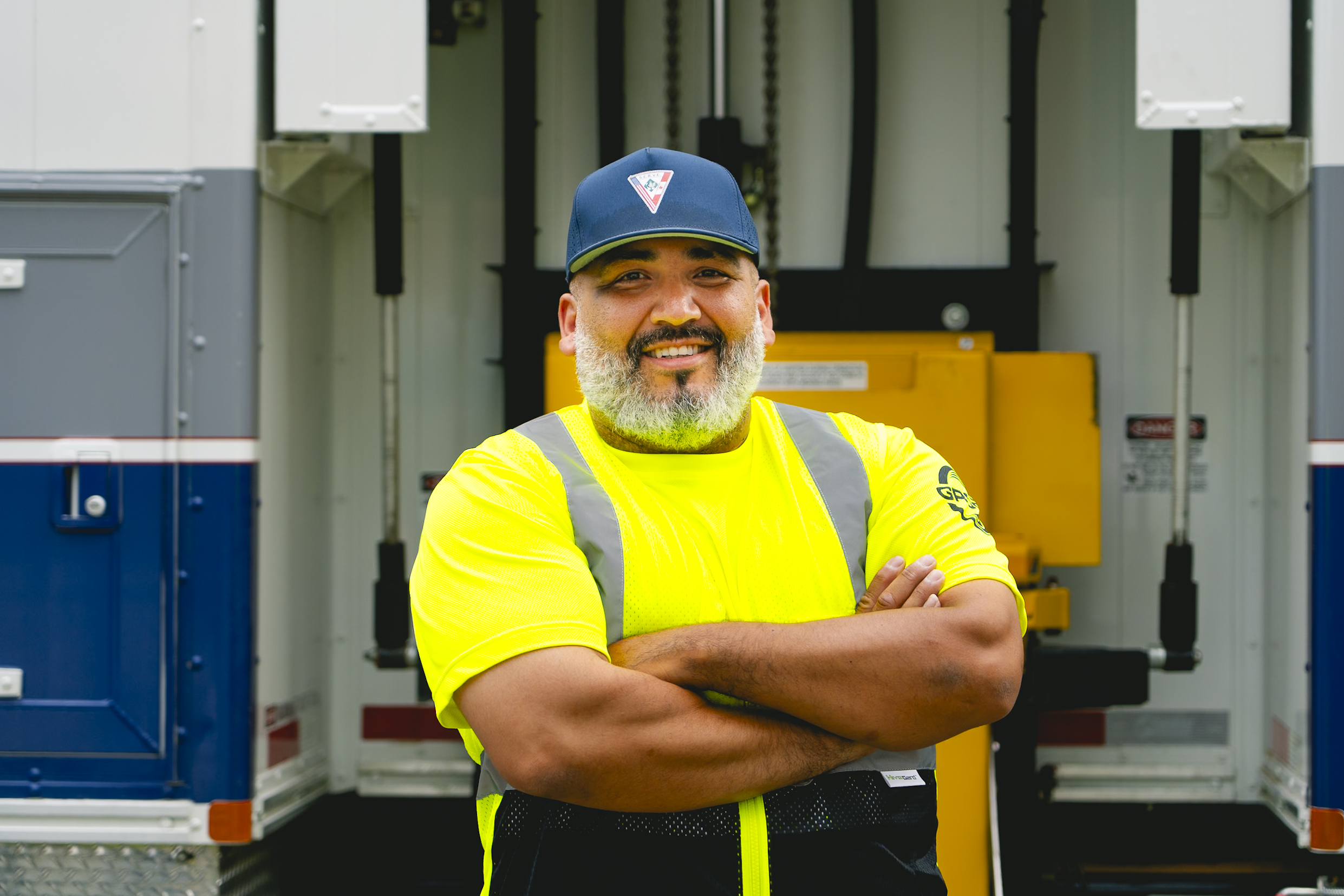 Smiling male worker in a neon yellow safety vest and cap with arms crossed in front of a truck.