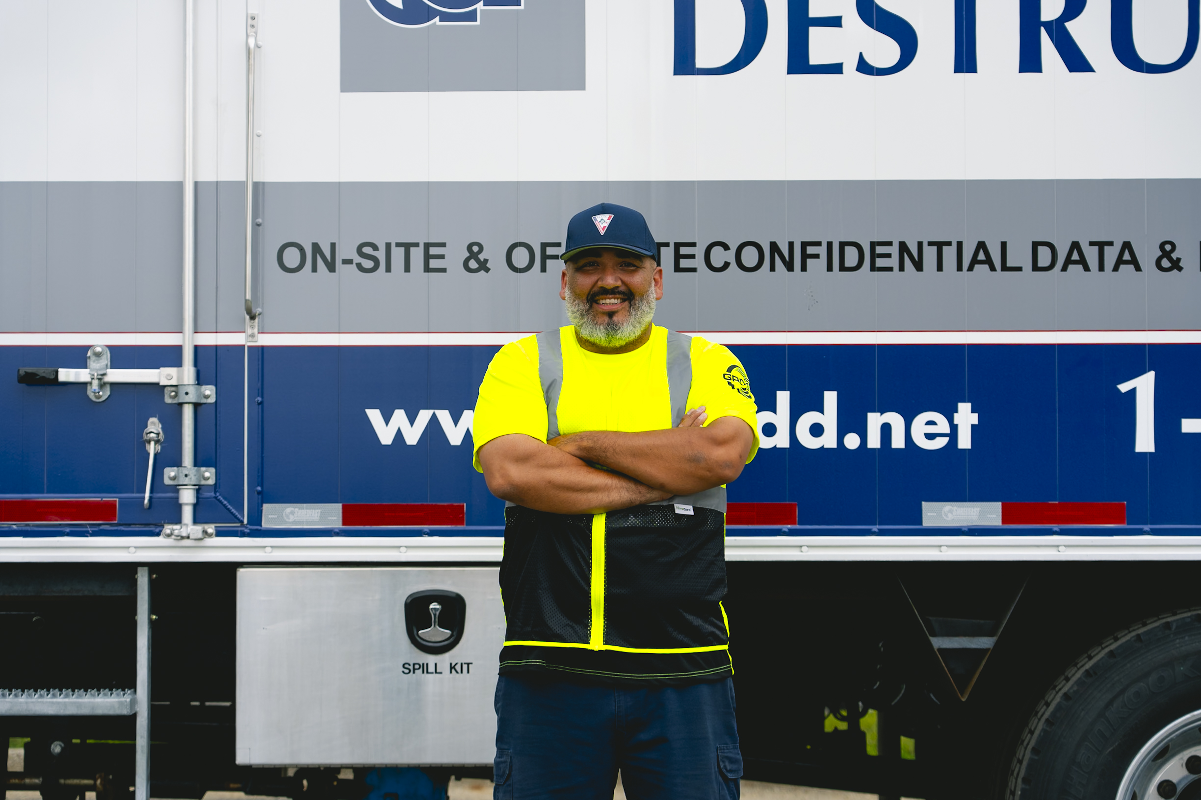 Man in yellow safety vest and cap standing with arms crossed in front of a work truck.