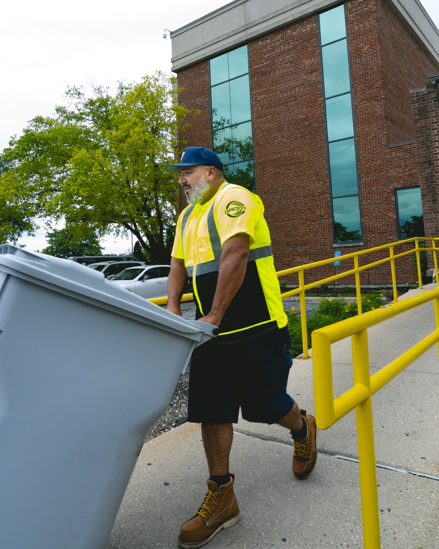 Man in yellow safety vest and boots pushing a large gray recycling bin near a brick building.