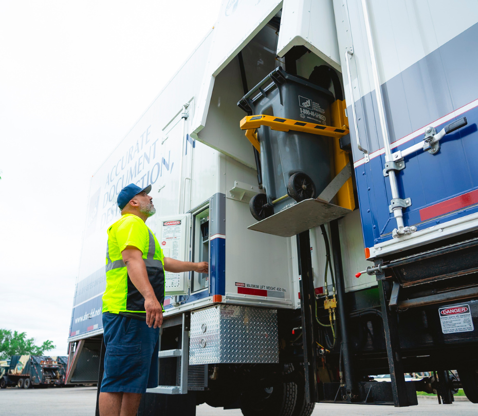 Man in neon shirt operating machine lifting a black shred bin into a white and blue truck.