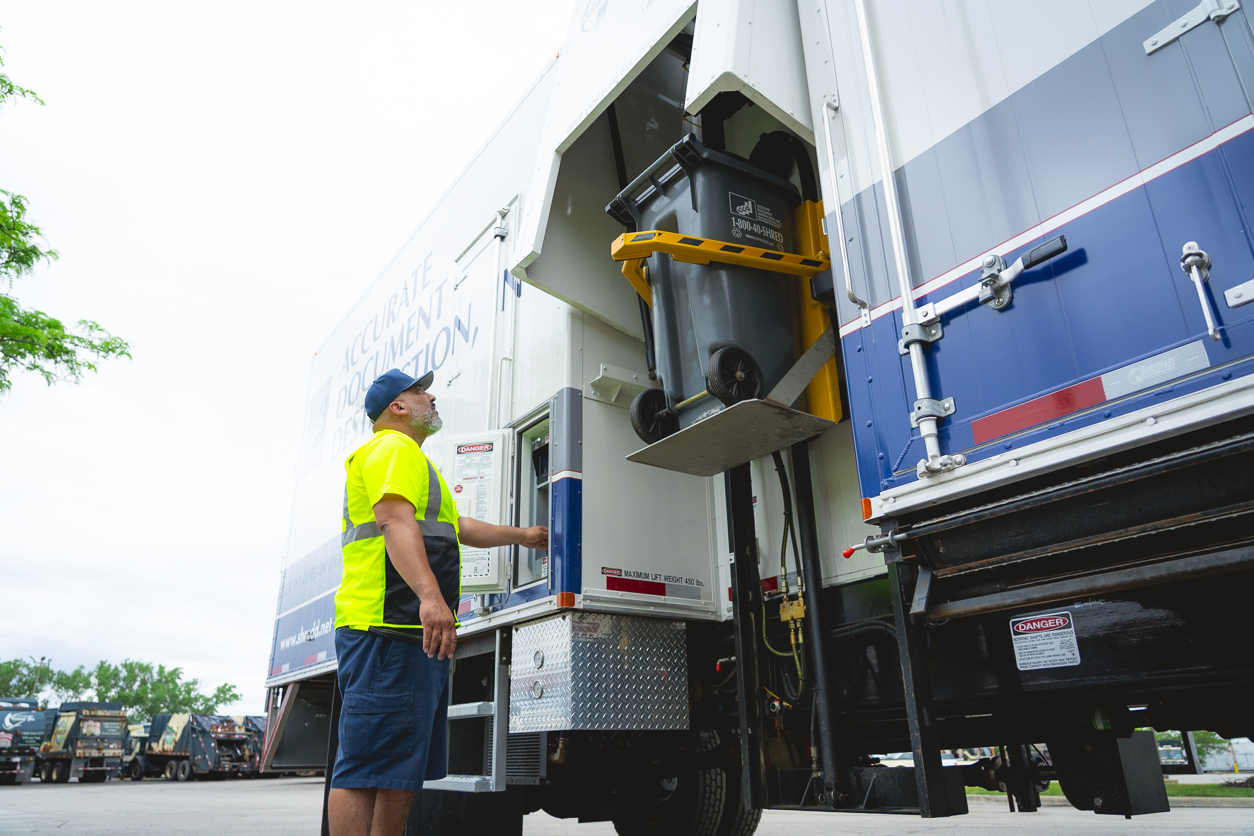 Worker in yellow shirt operating a waste bin lift mechanism on a large white and blue truck.