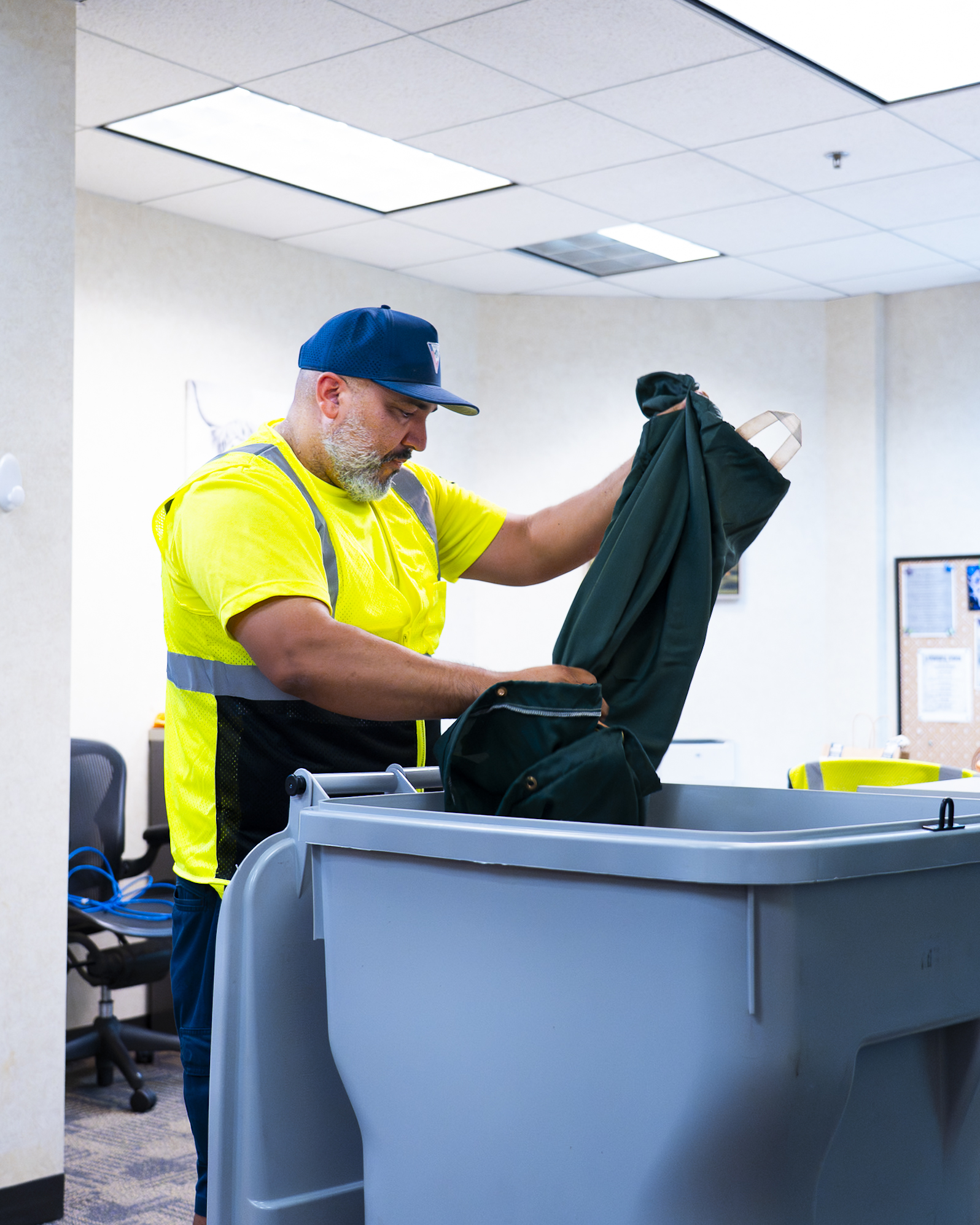 Worker in yellow safety vest and blue cap removes green coat from large gray plastic bin indoors.