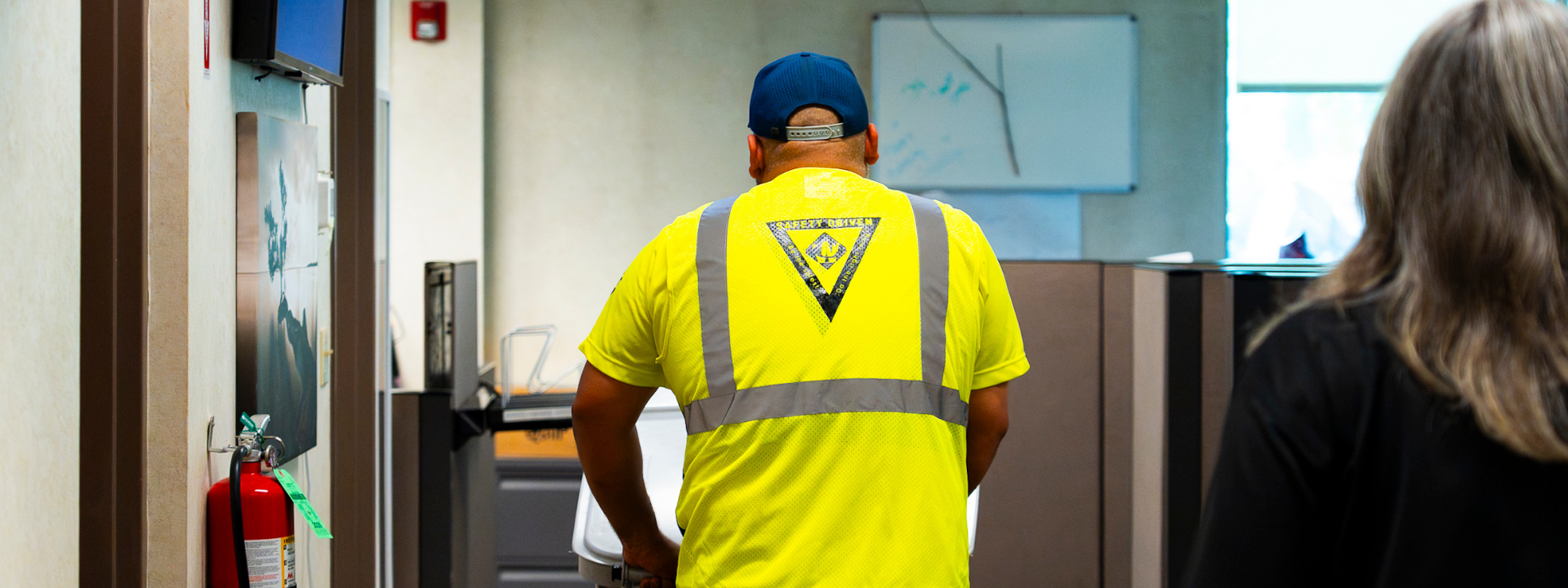 Man in a yellow safety vest and blue cap seen from behind inside an office hallway.