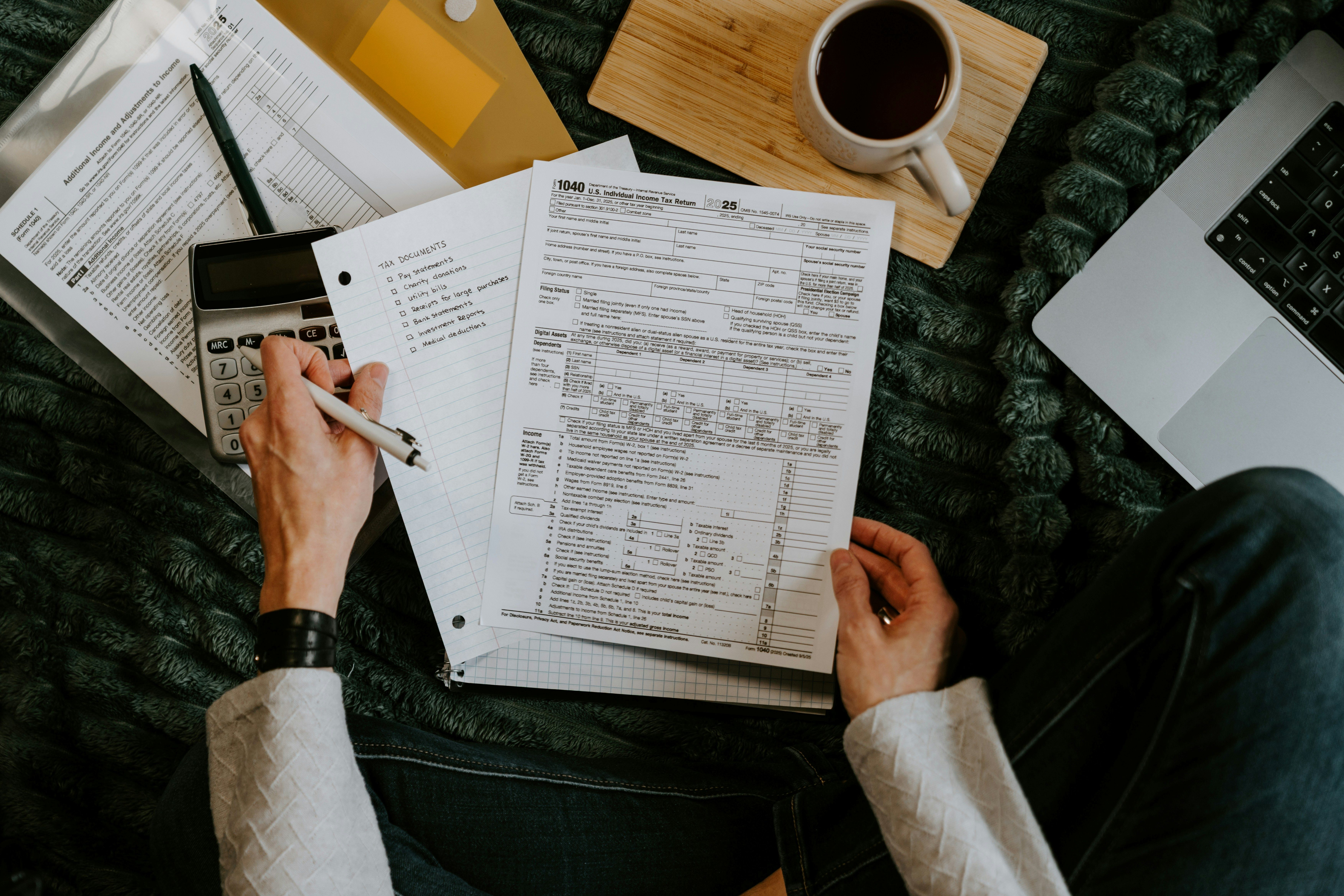 Person filling out a 2025 U.S. tax form 1040 with calculator, checklist, coffee, and laptop nearby.