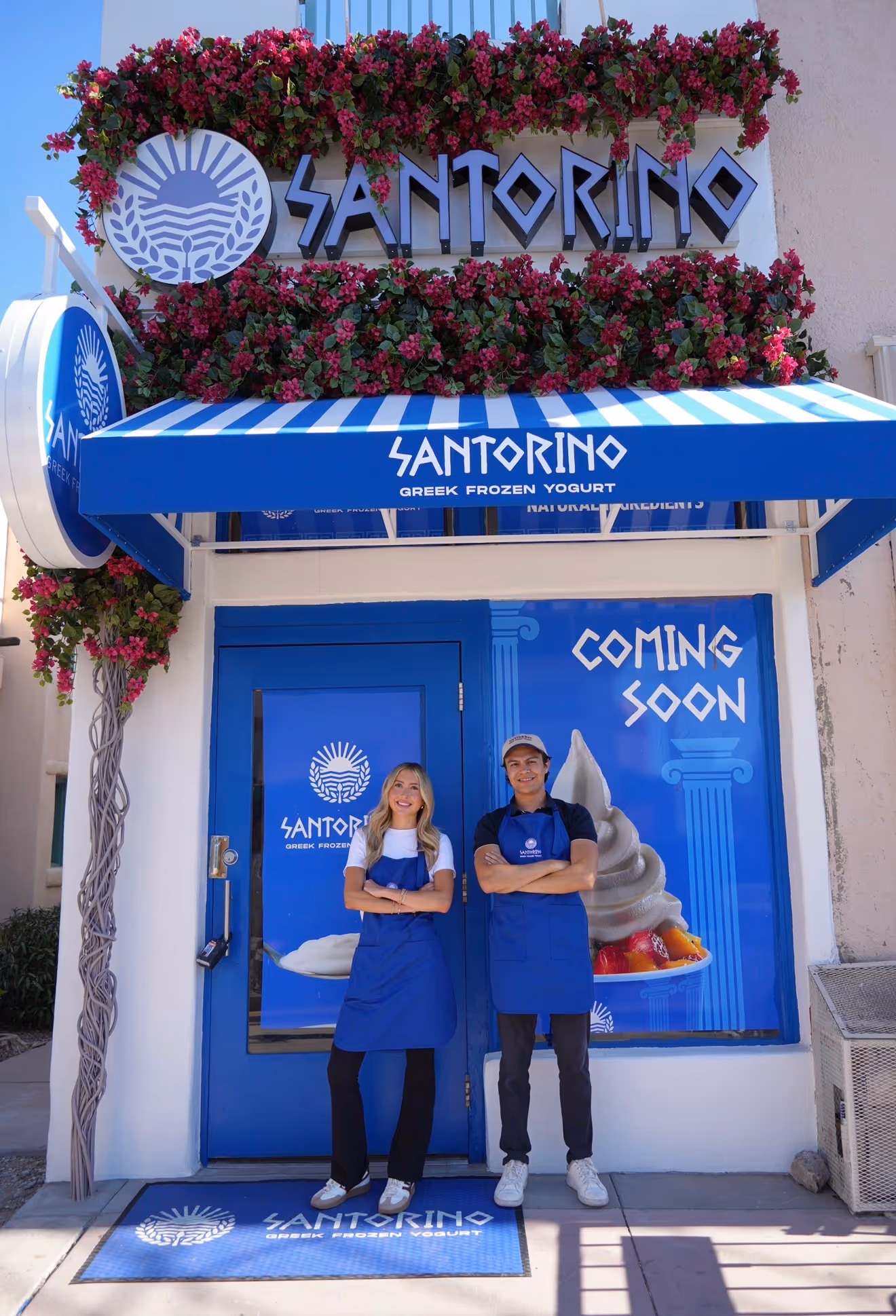 Two Santorino employees standing in blue aprons outside a Greek frozen yogurt shop with a 'Coming Soon' sign.
