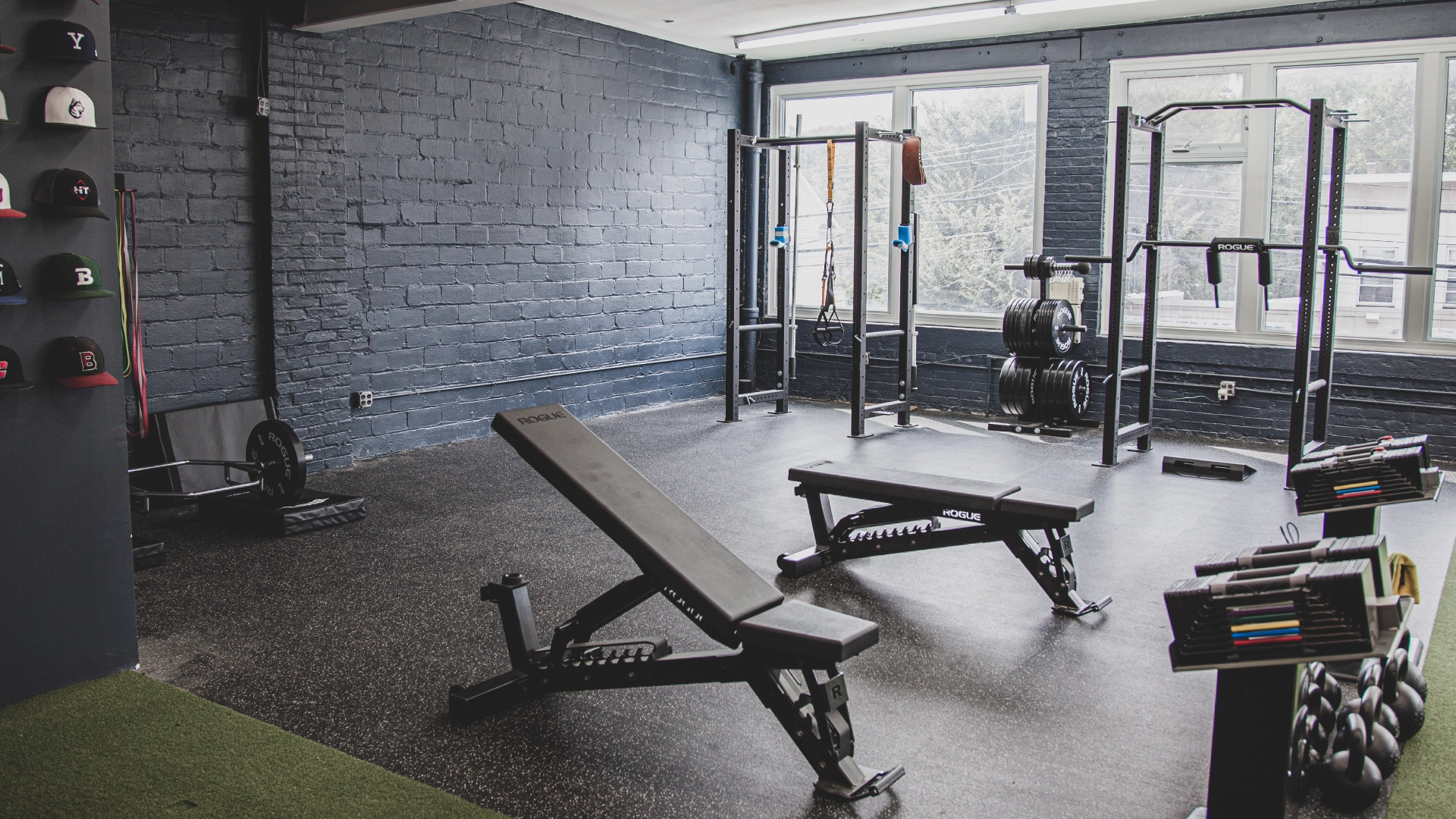 Minimalist gym with black benches, squat racks, and weights on dark flooring. Wall-mounted hats and large windows create a clean, inviting atmosphere.
