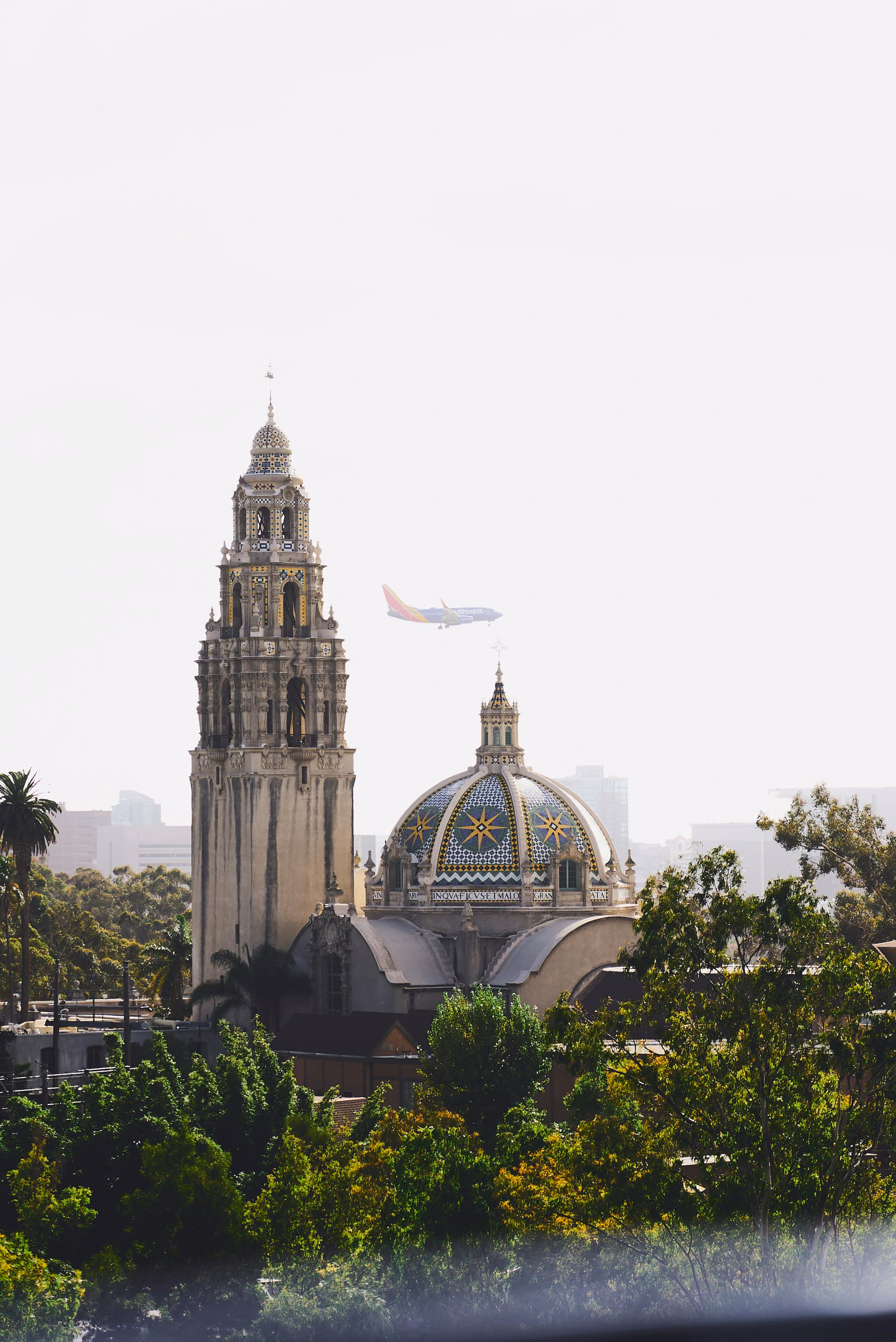 Historic tower and dome with decorative tiles rising above green trees, with a Southwest airplane flying in the background.